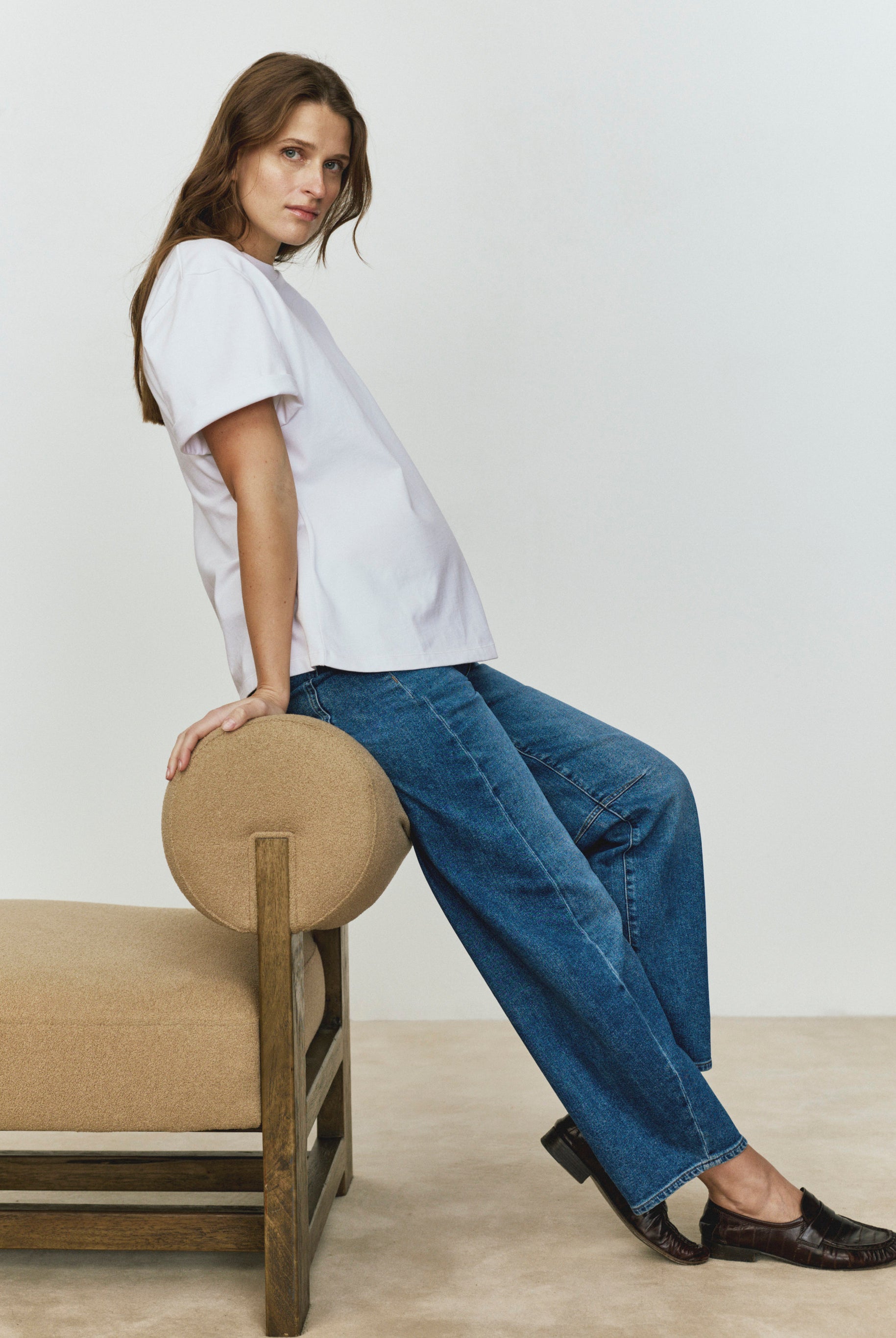 Woman wearing a white t-shirt and blue jeans sitting on a beige chair against a plain background
