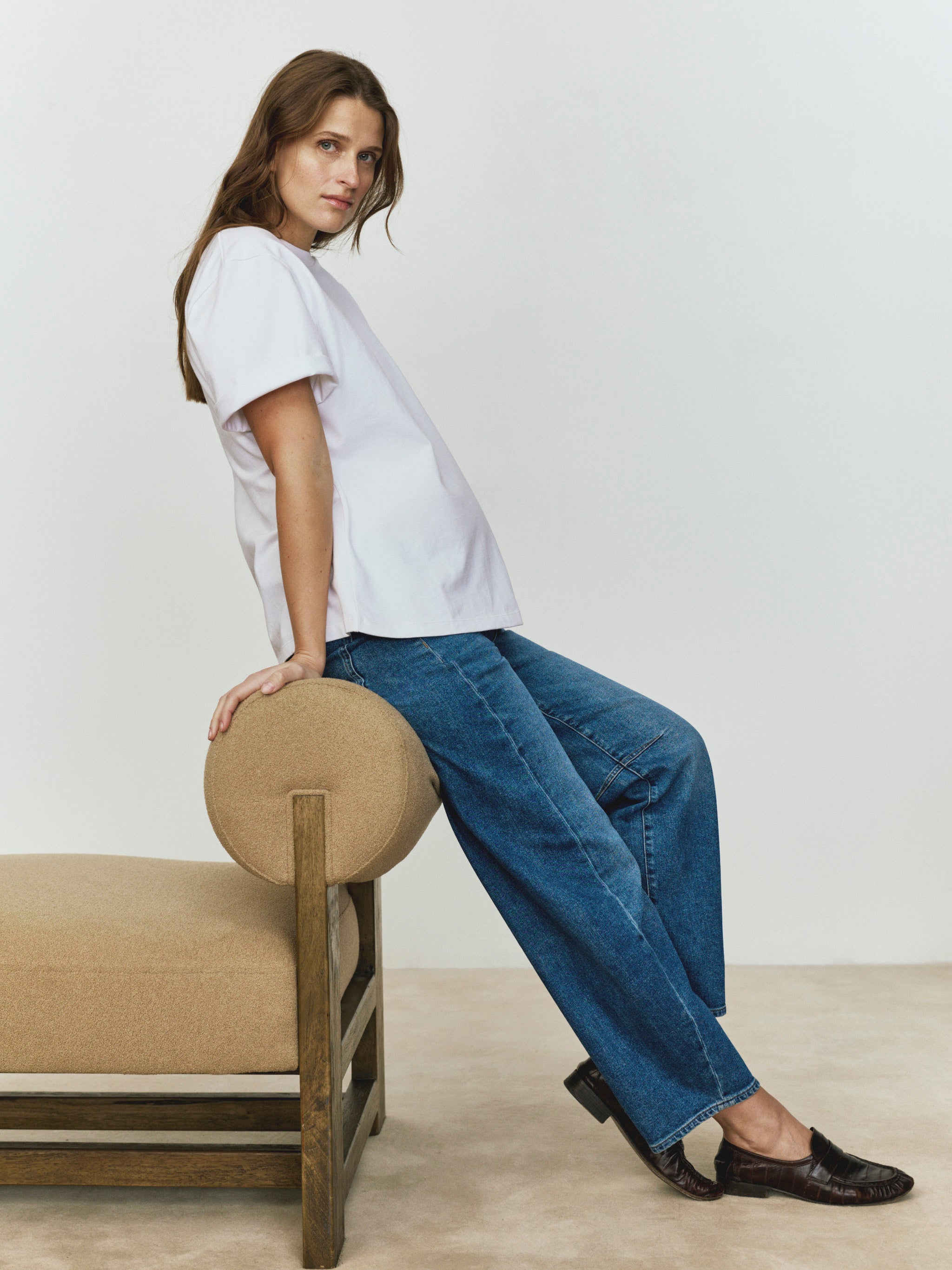 Woman wearing a white t-shirt and blue jeans sitting on a beige chair against a plain background