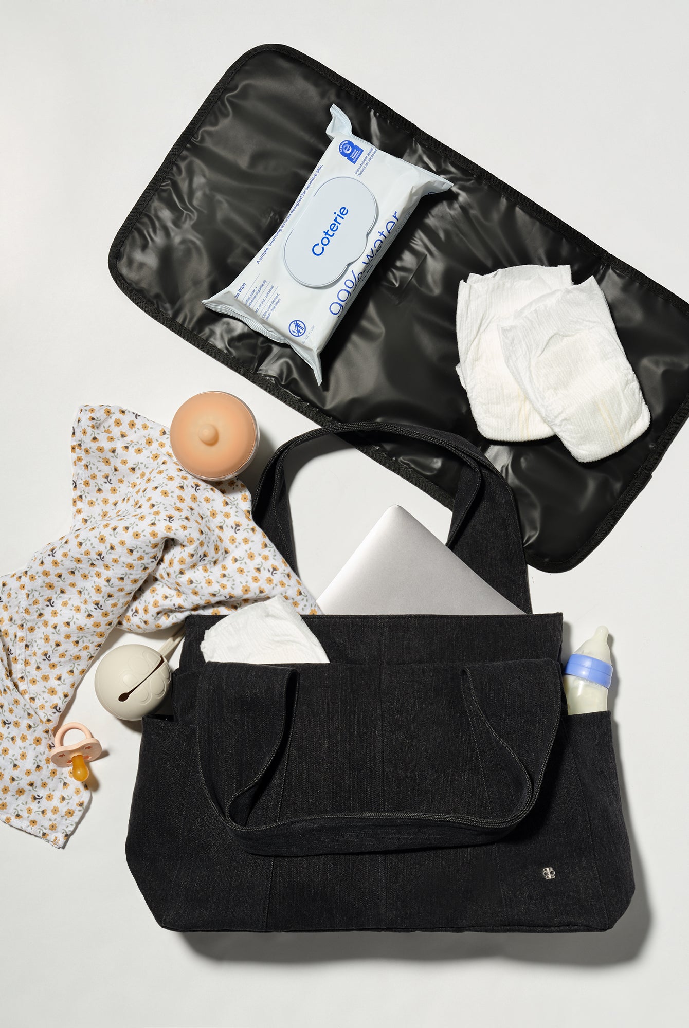 A black diaper bag shown open in a flat-lay arrangement, filled with baby essentials including a patterned baby shirt, wipes, diapers, a pacifier, and a bottle, on a light gray background.