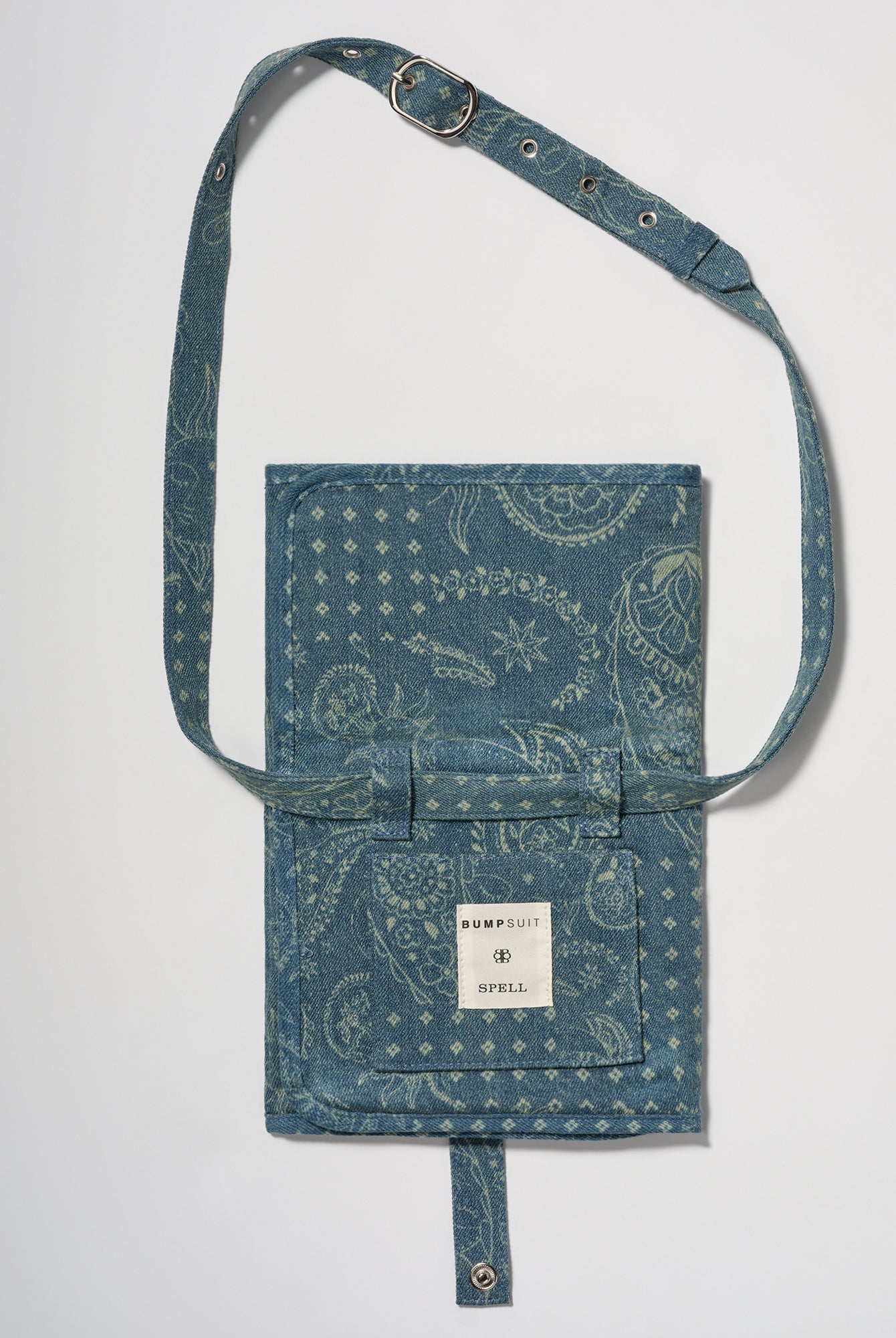 A blue denim crossbody diaper change mat bag by with a paisley and geometric pattern by Spell and Bumpsuit,, shown on a light gray background.
