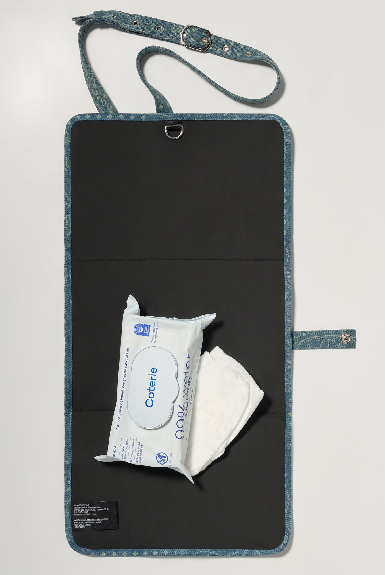 A denim roll up diaper change mat bag by Spell and Bumpsuit with a black inside lining, shown on a light gray background with wipes and diapers