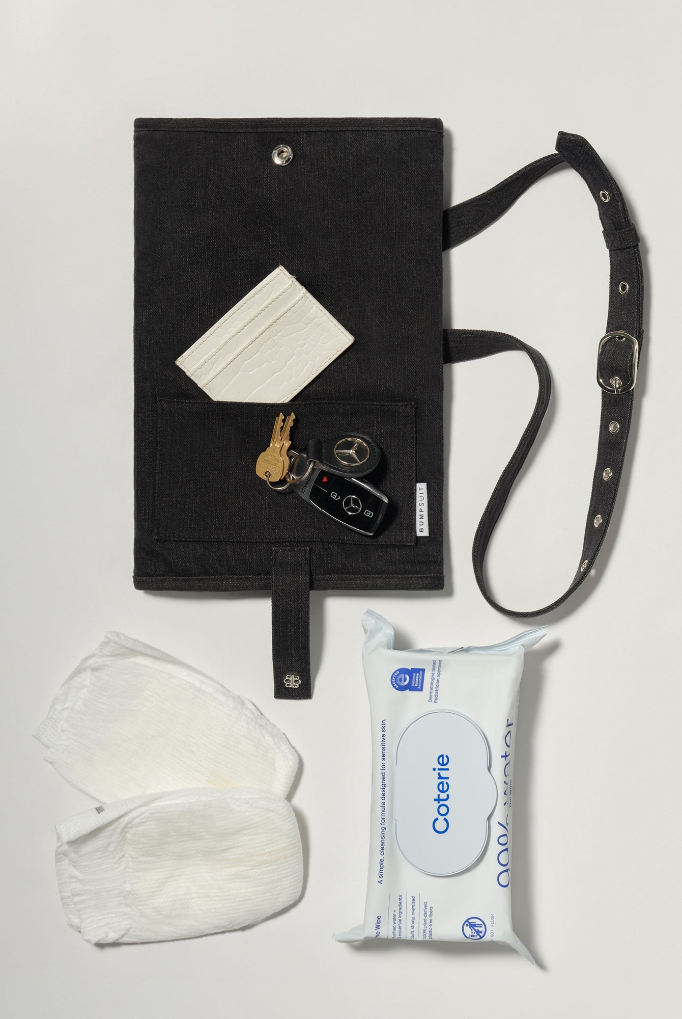 A black denim roll up diaper change mat bag by Spell and Bumpsuit, shown on a light gray background with wipes, diapers, keys, and wallet