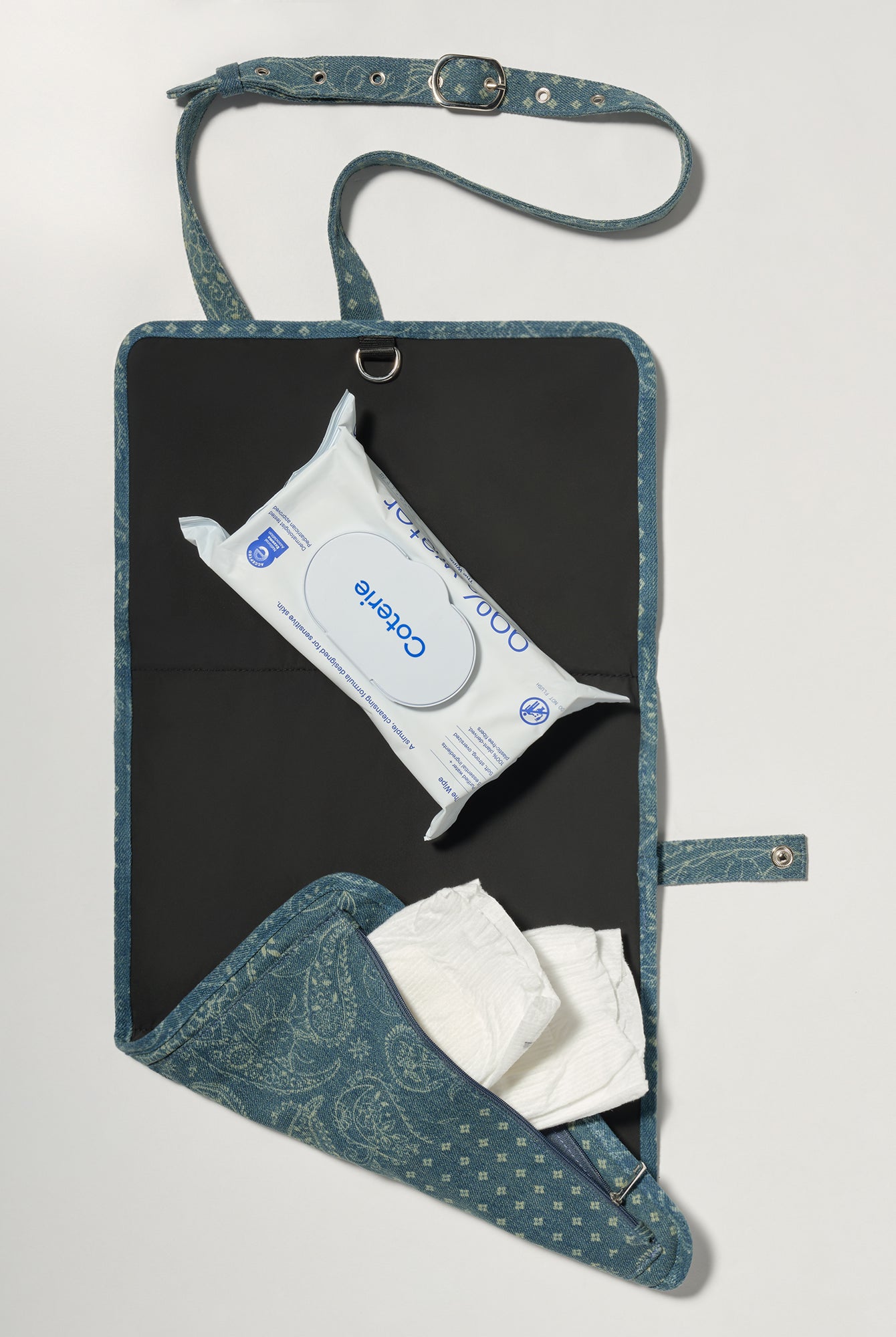A blue patterned diaper wrap bag with a black interior, shown open on a light gray background. It contains a pack of wipes and white diapers.