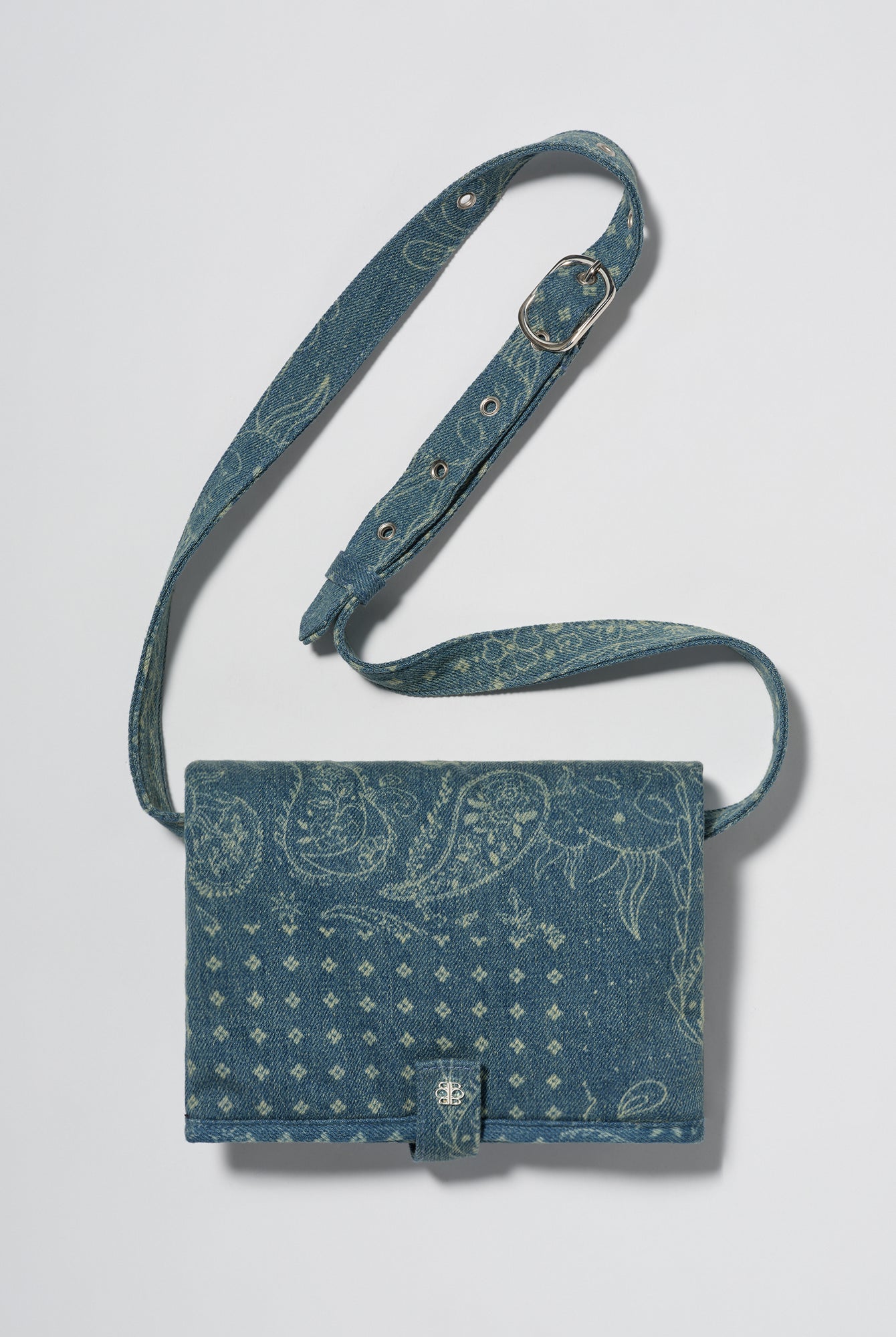 A blue denim crossbody diaper change mat bag with a paisley and geometric pattern by Spell and Bumpsuit,, shown on a light gray background.