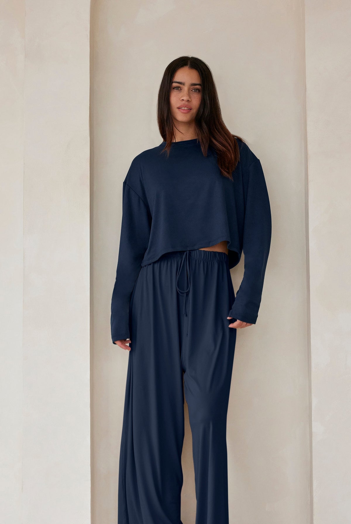 A woman standing against a beige feature wall wearing Bumpsuits Cloud lounge pants and a long sleeve crop tee in navy