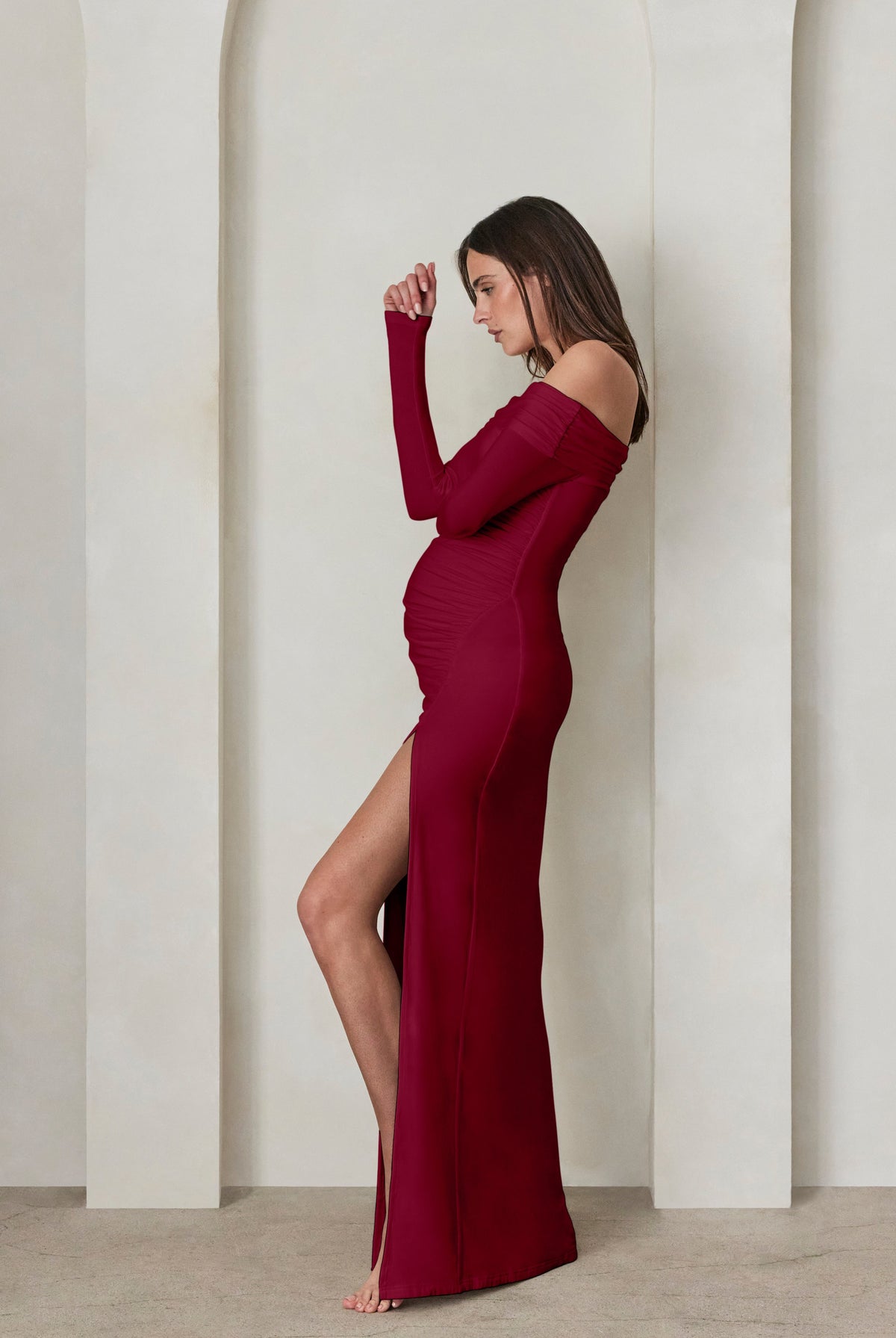 A pregnant woman wearing a Bumpsuit burgundy off the shoulder maxi dress with high leg slit against a beige feature wall