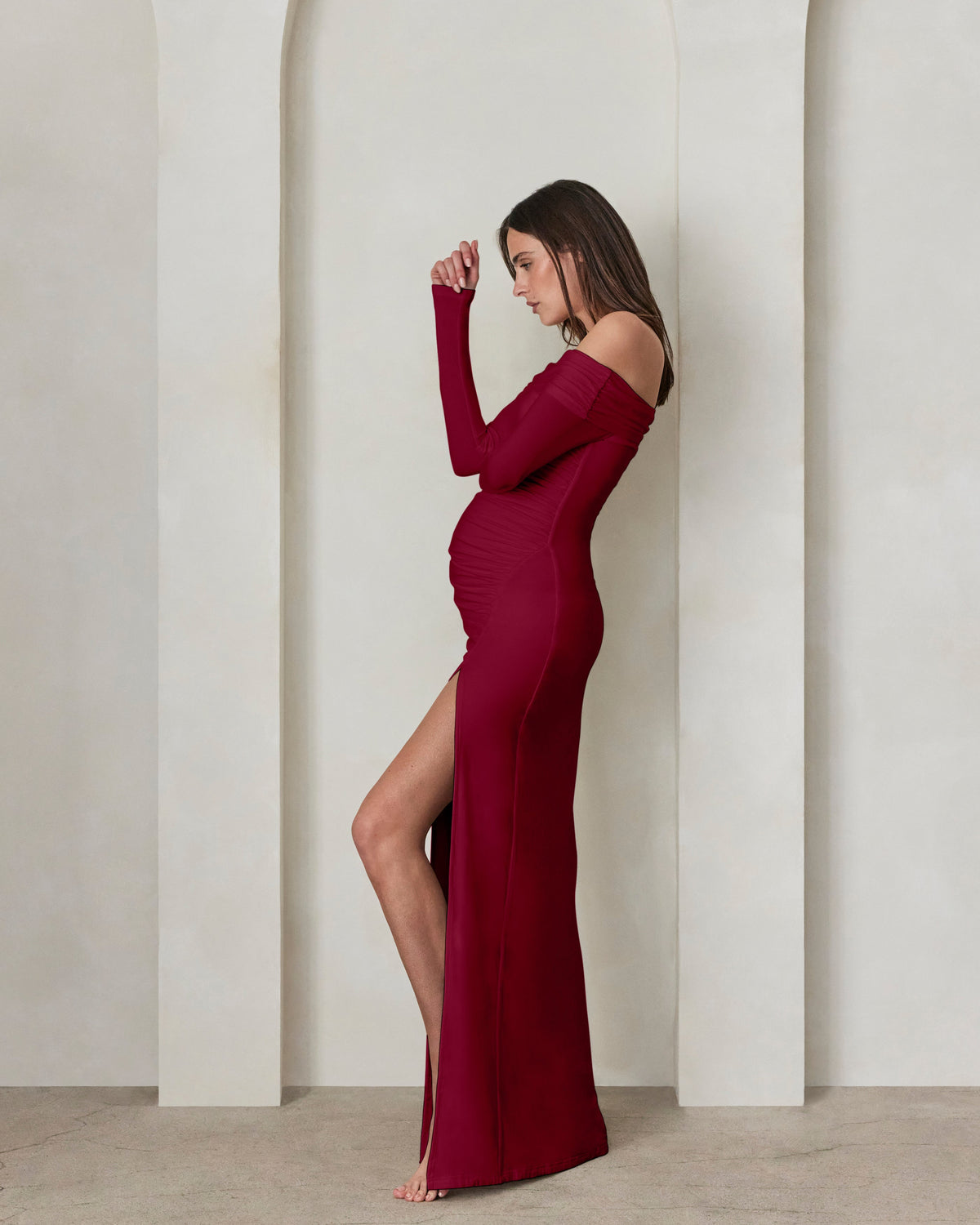 A pregnant woman wearing a Bumpsuit burgundy off the shoulder maxi dress with high leg slit against a beige feature wall
