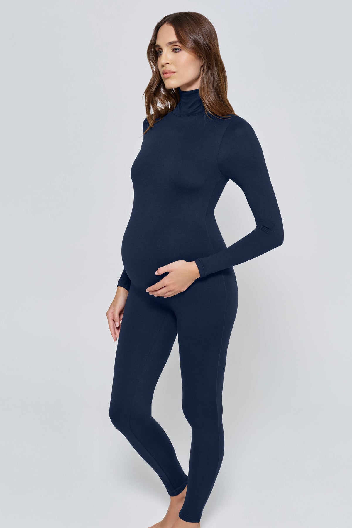 A pregnant woman modeling a dark navy blue maternity turtleneck jumpsuit against a light gray studio background.
