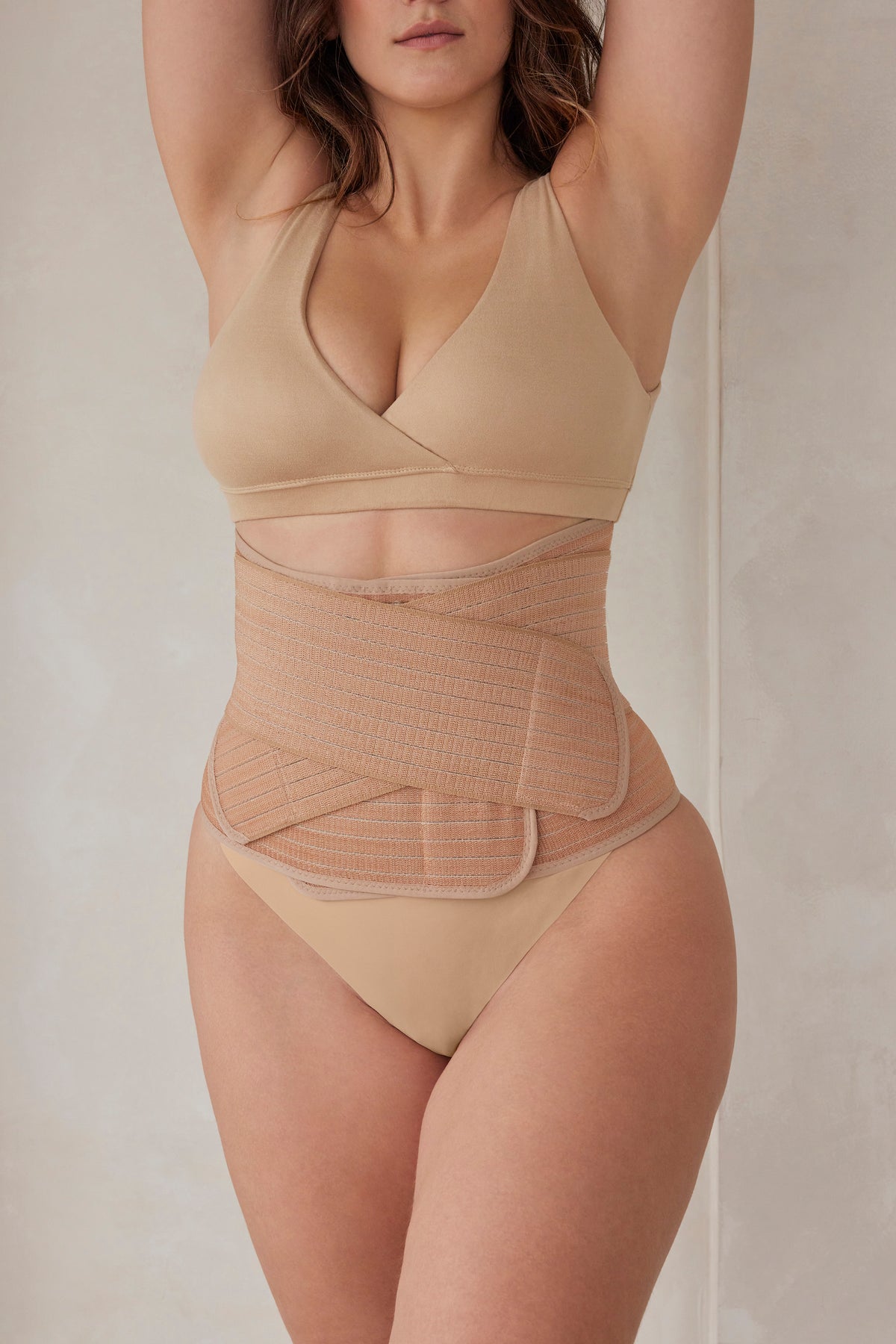 Pregnant woman wearing a beige reversible comfy bra and velcro waist trainer against a plain background
