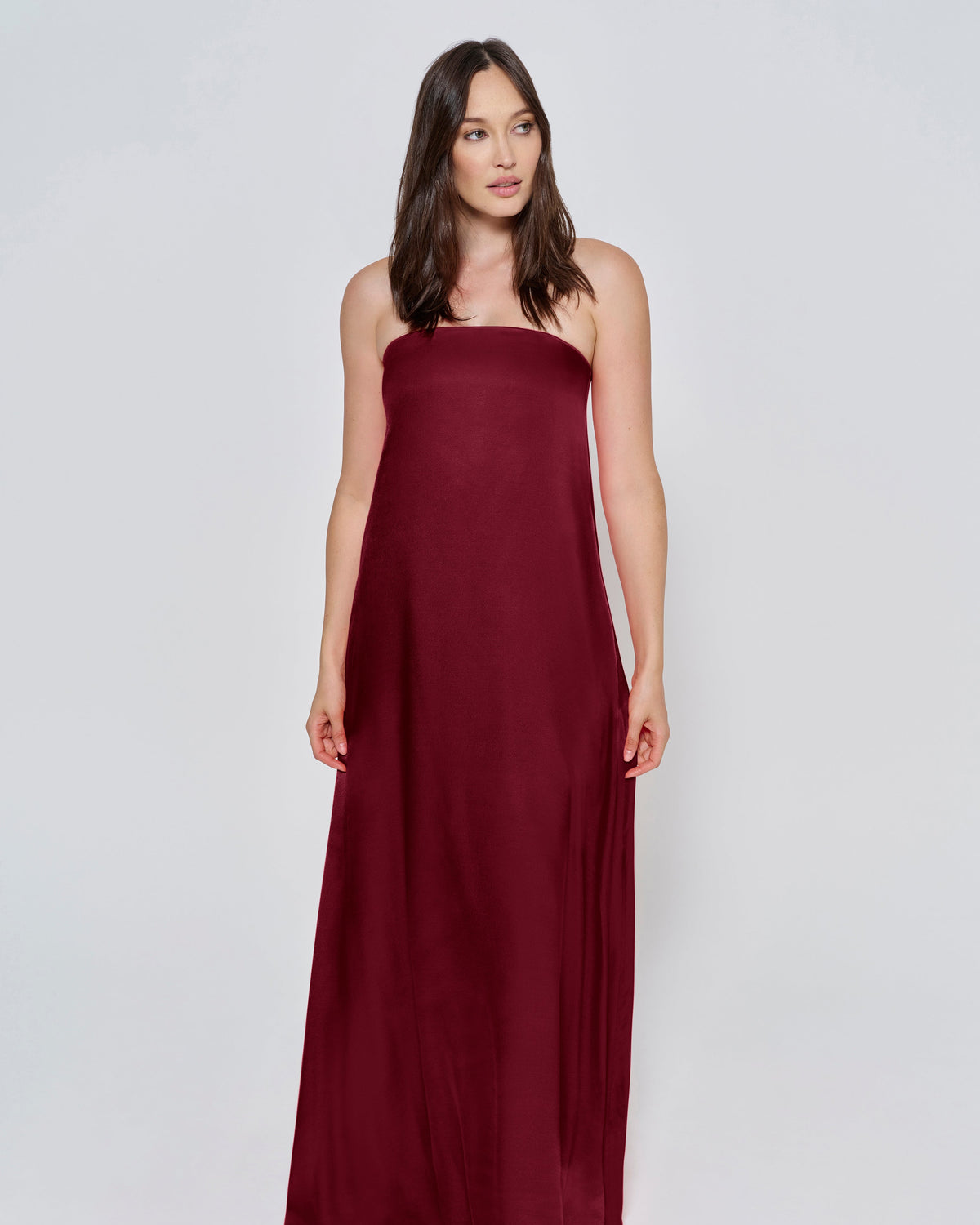Pregnant woman wearing a Bumpsuit brushed satin gown in a Burgundy color against a white background