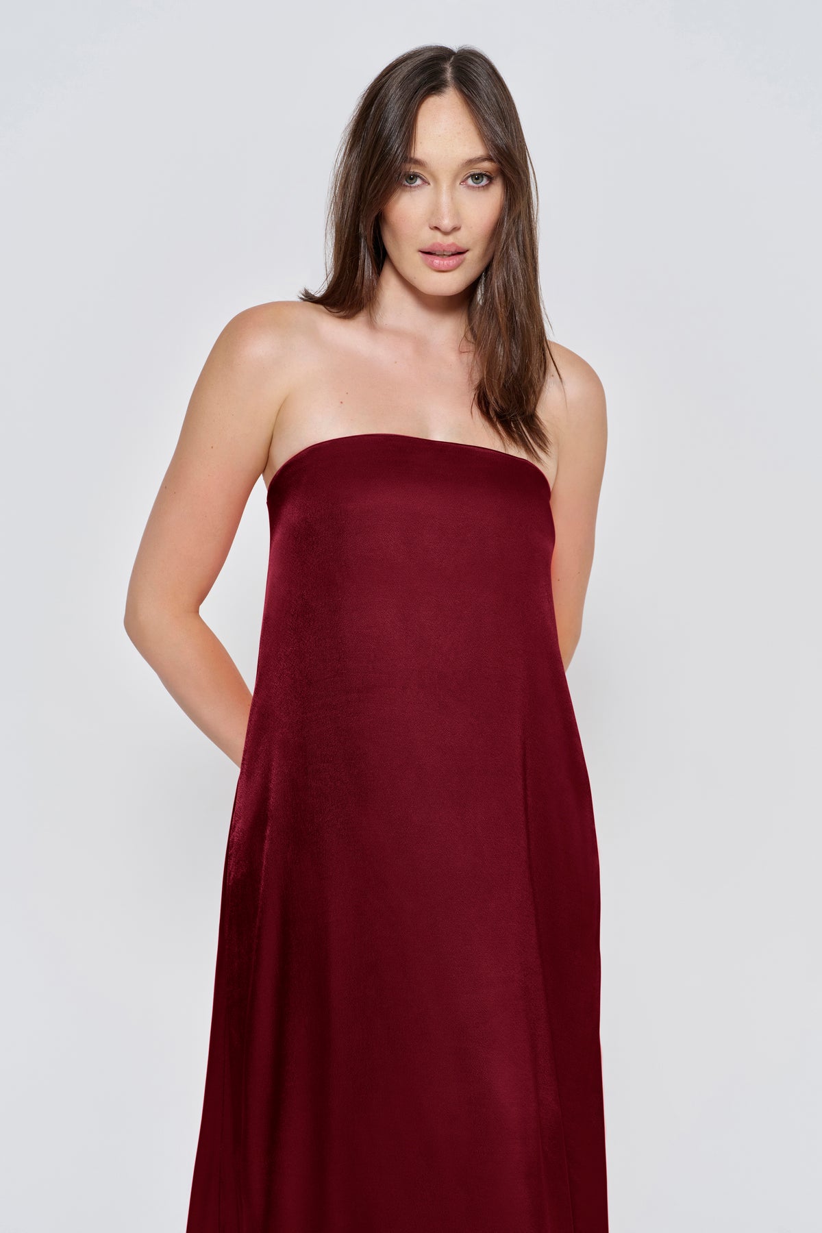 Pregnant woman wearing a Bumpsuit brushed satin gown in a Burgundy color against a white background