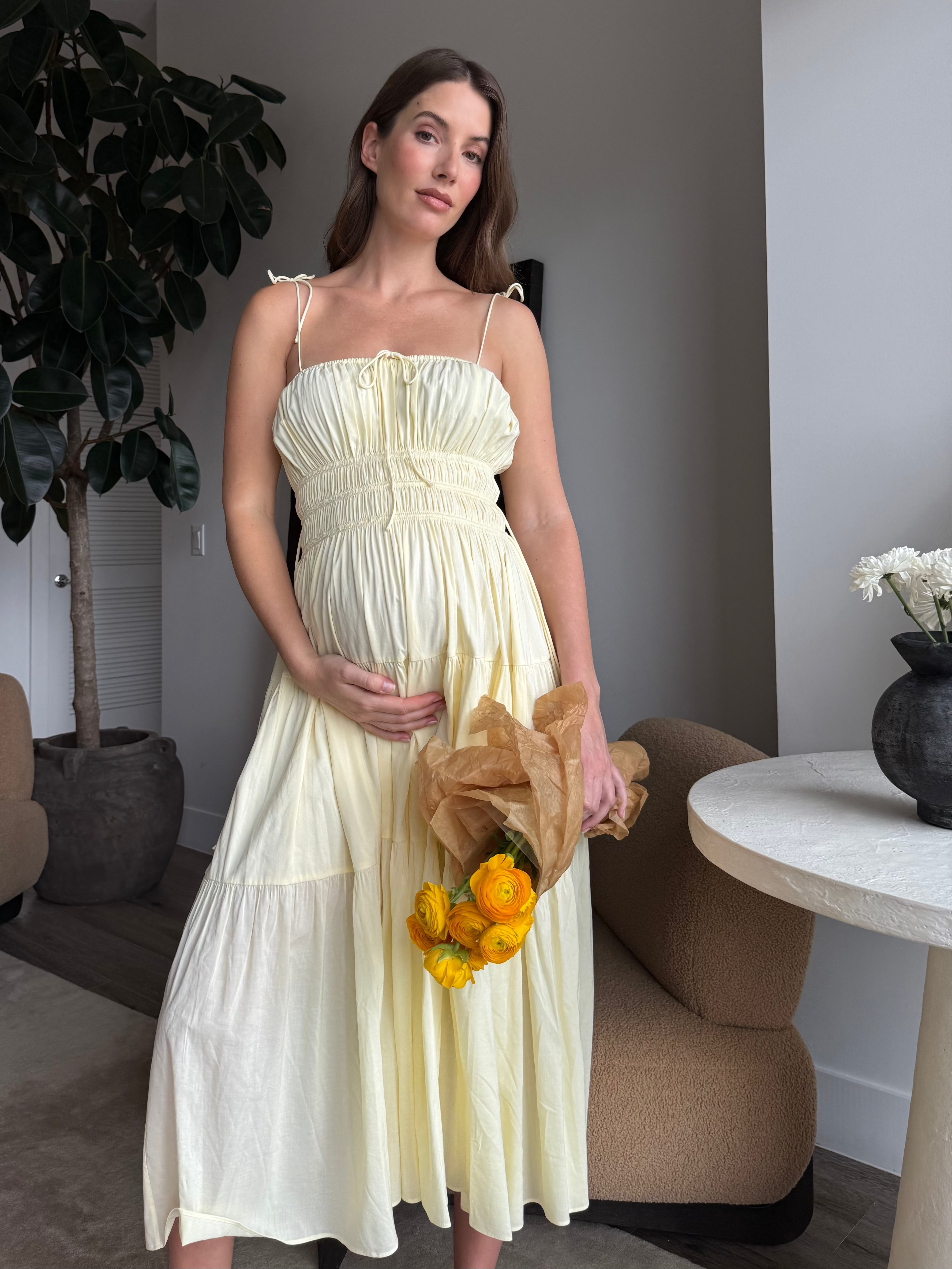 Woman in a light yellow shirred dress holding flowers in a room with a plant and table.