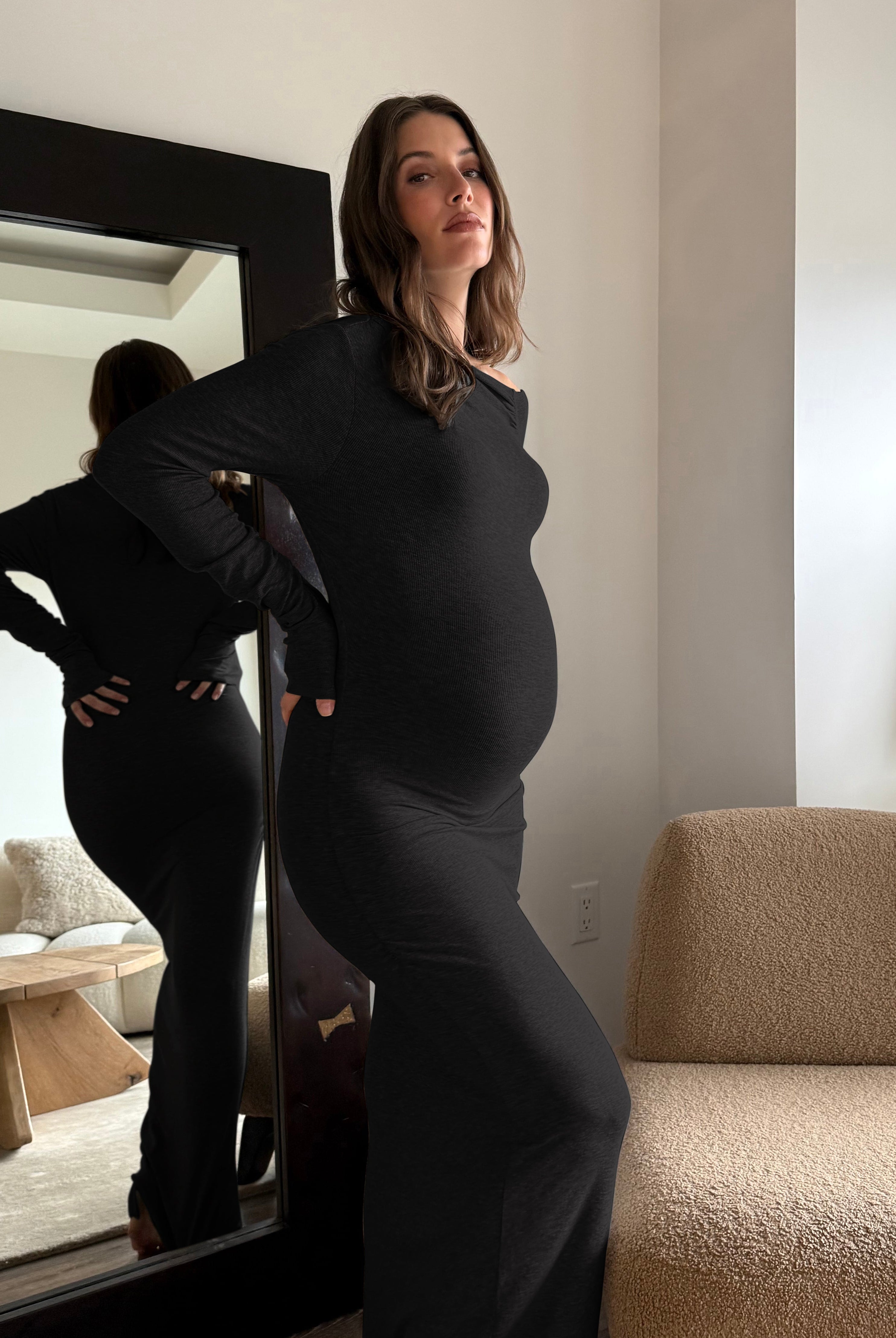 A pregnant woman wearing a fitted black long-sleeve maternity dress stands indoors next to a mirror that reflects her back. The room has white walls, a beige boucle chair, and a side table with a vase.