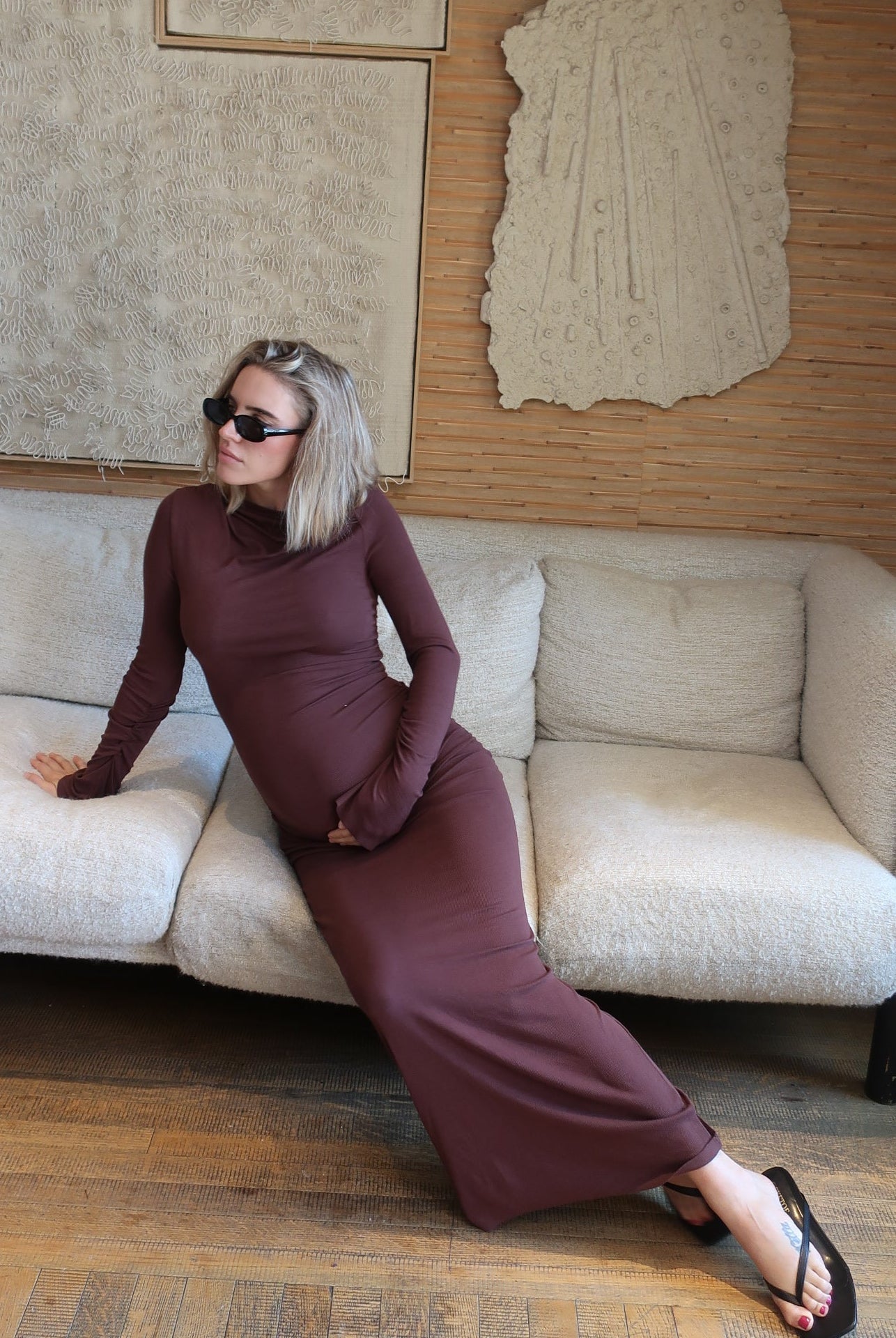A pregnant woman wearing a soft rib maxi dress in chocolate brown by Bumpsuit, sitting on a beige sofa