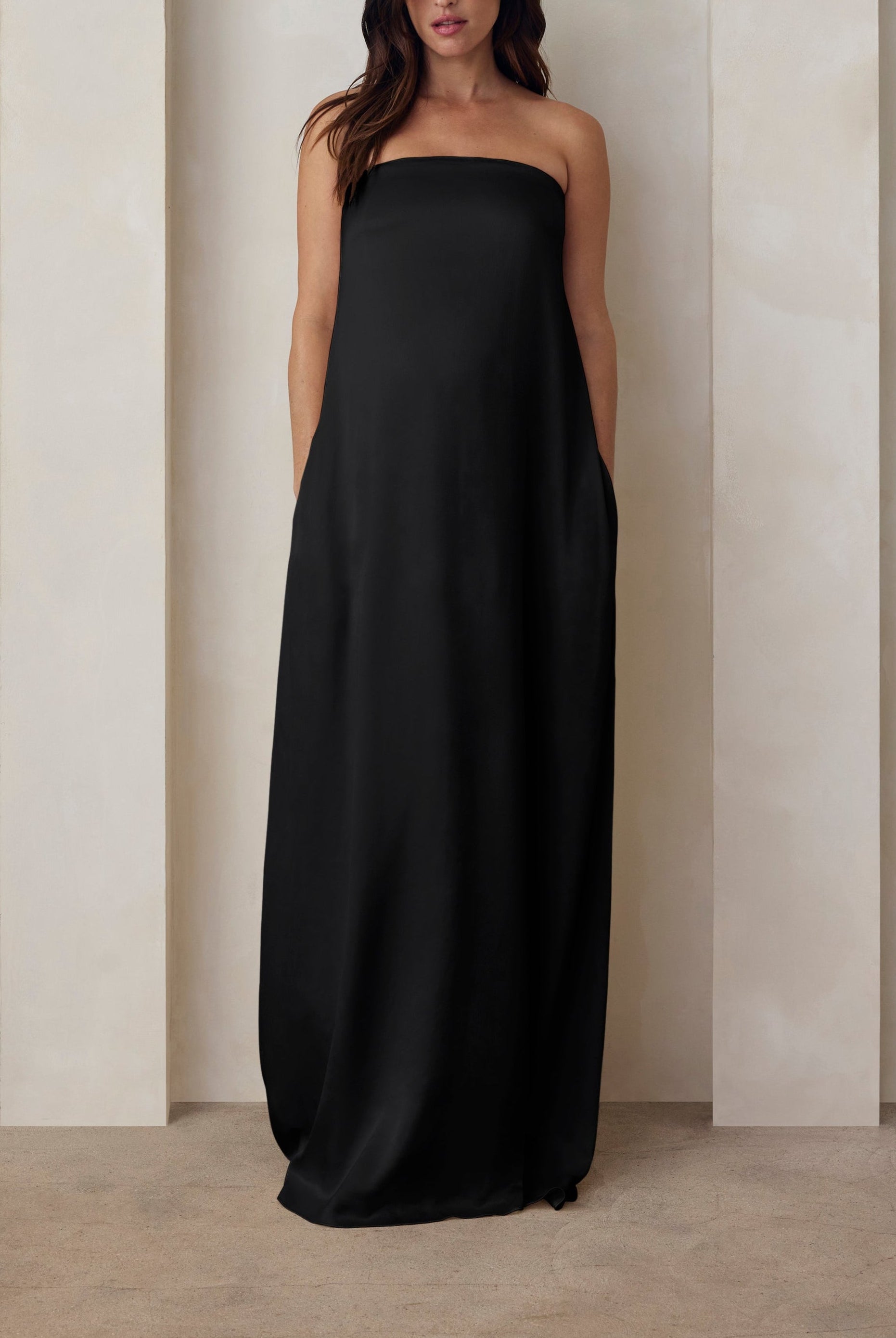 A pregnant woman wearing the Bumpsuit brushed satin strapless maxi maternity gown in black against a beige feature wall