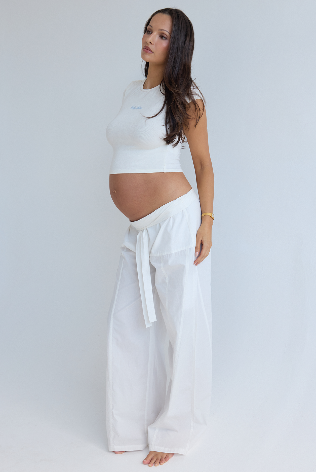 A pregnant woman stands in profile wearing a white sleeveless crop top and matching white wide-leg pants by Bumpsuit, photographed against a plain light gray studio background.