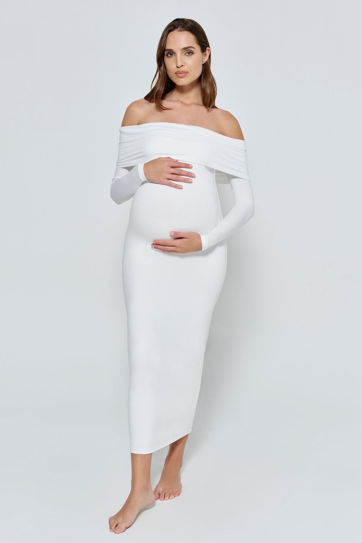 Pregnant woman wearing a ivory off-shoulder dress against a white background