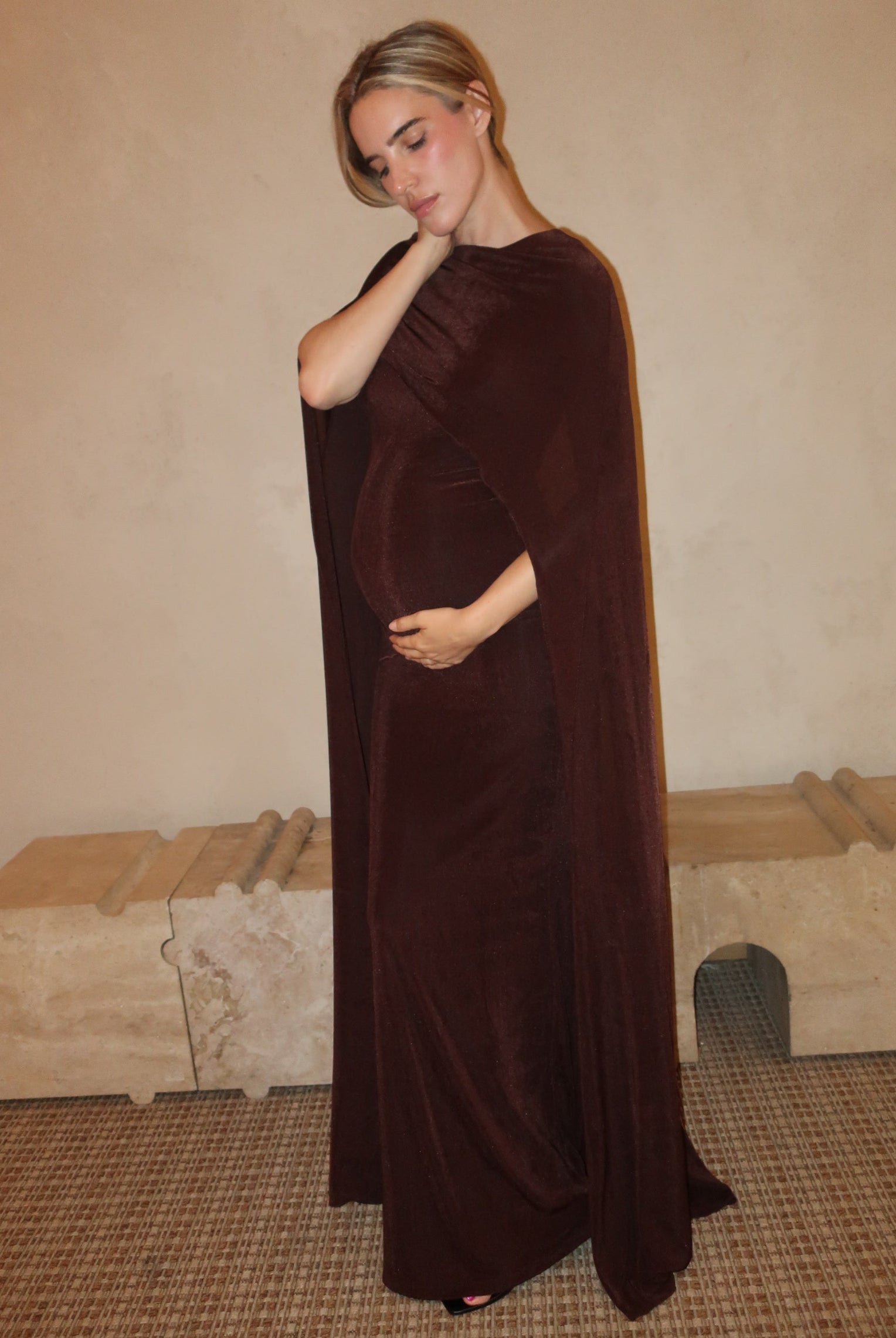 A pregnant woman wearing a Bumpsuit maxi dress with a floor length cape detail in chocolate brown against a neutral setting