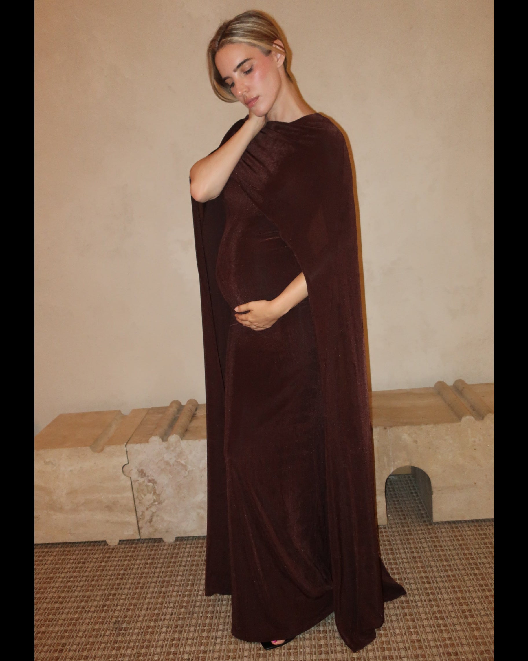 A pregnant woman wearing a Bumpsuit maxi dress with a floor length cape detail in chocolate brown against a neutral setting