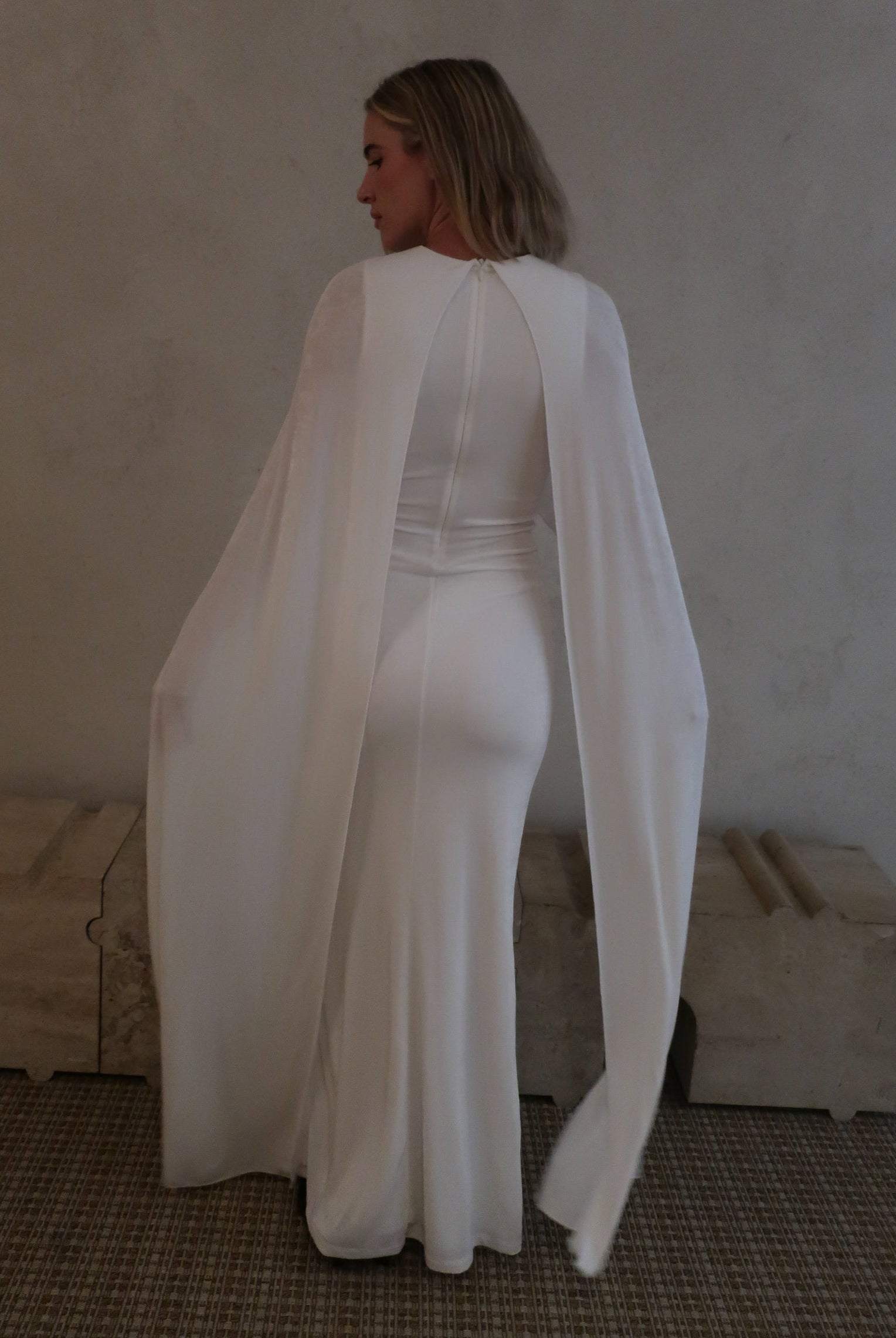 A woman wearing Bumpsuit maxi gown with a long sheer cape, standing in a neutral indoor setting.