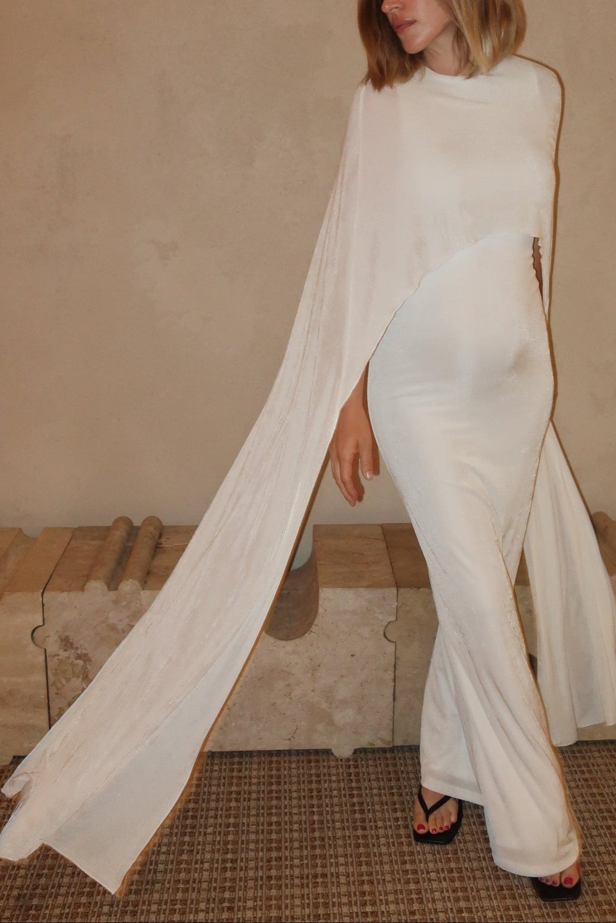 A pregnant woman wearing a Bumpsuit maxi cape dress in ivory against a beige background