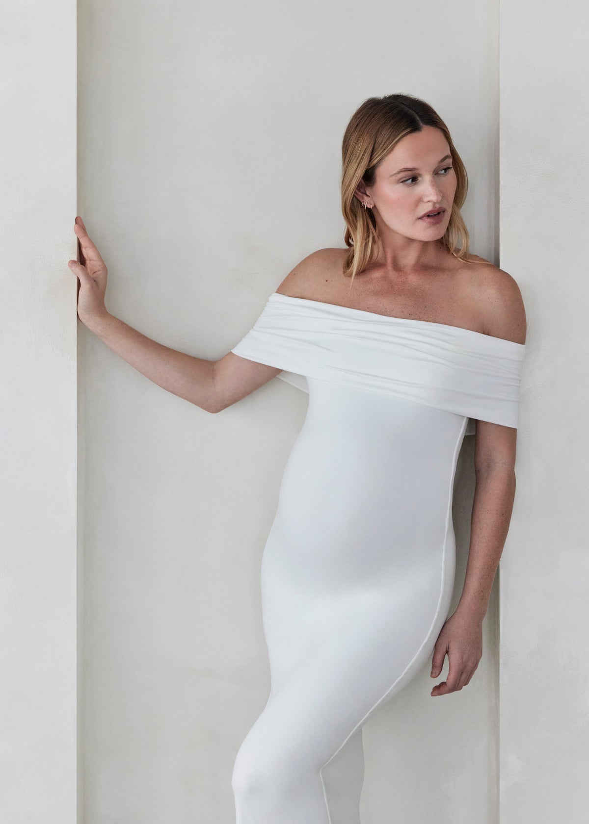 Pregnant woman wearing Bumpsuits off the shoulder Ivory Bianca maternity maxi dress standing against a beige wall
