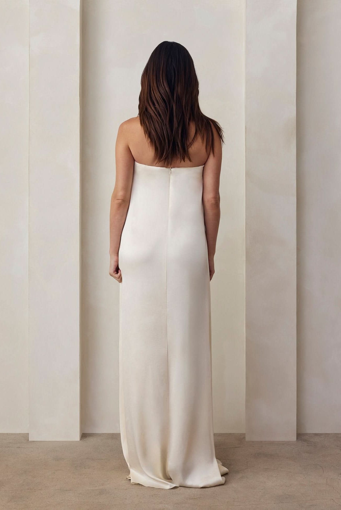 the back view of a pregnant model wearing the Bumpsuit brushed satin gown in ivory against a beige feature wall
