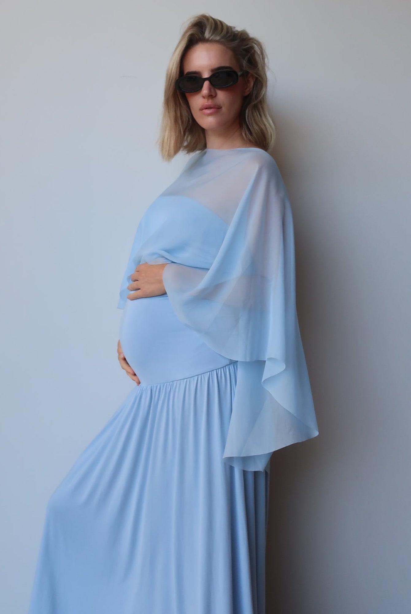A pregnant woman wearing a light blue maternity dress with a sheer chiffon cape by bumpsuit and sunglasses, posing against a plain light gray wall.