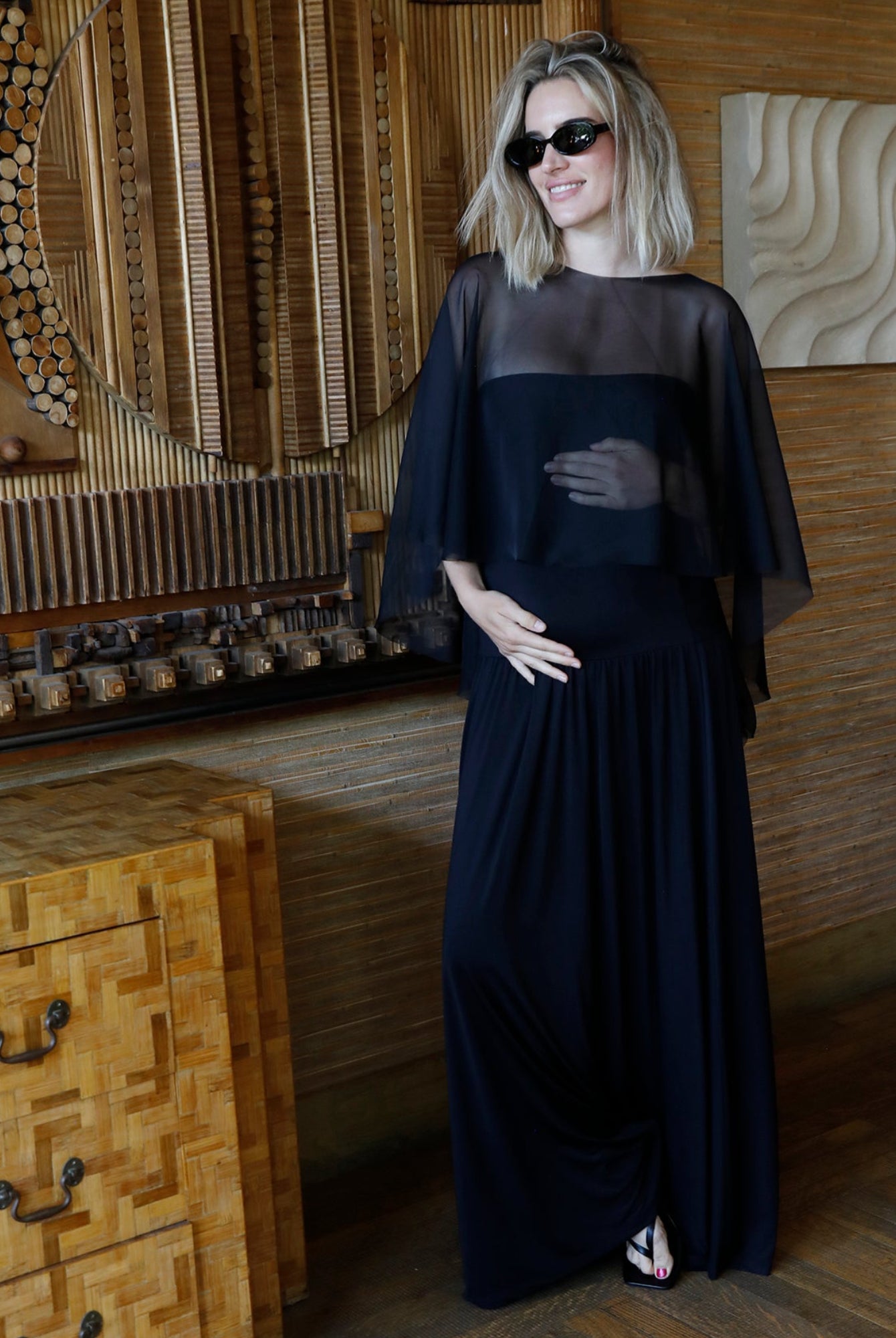 A woman wearing Bumpsuits Maria dress and chiffon Capelet in black, styled with black sunglasses, standing in a rustic room with wooden furniture and wall art.
