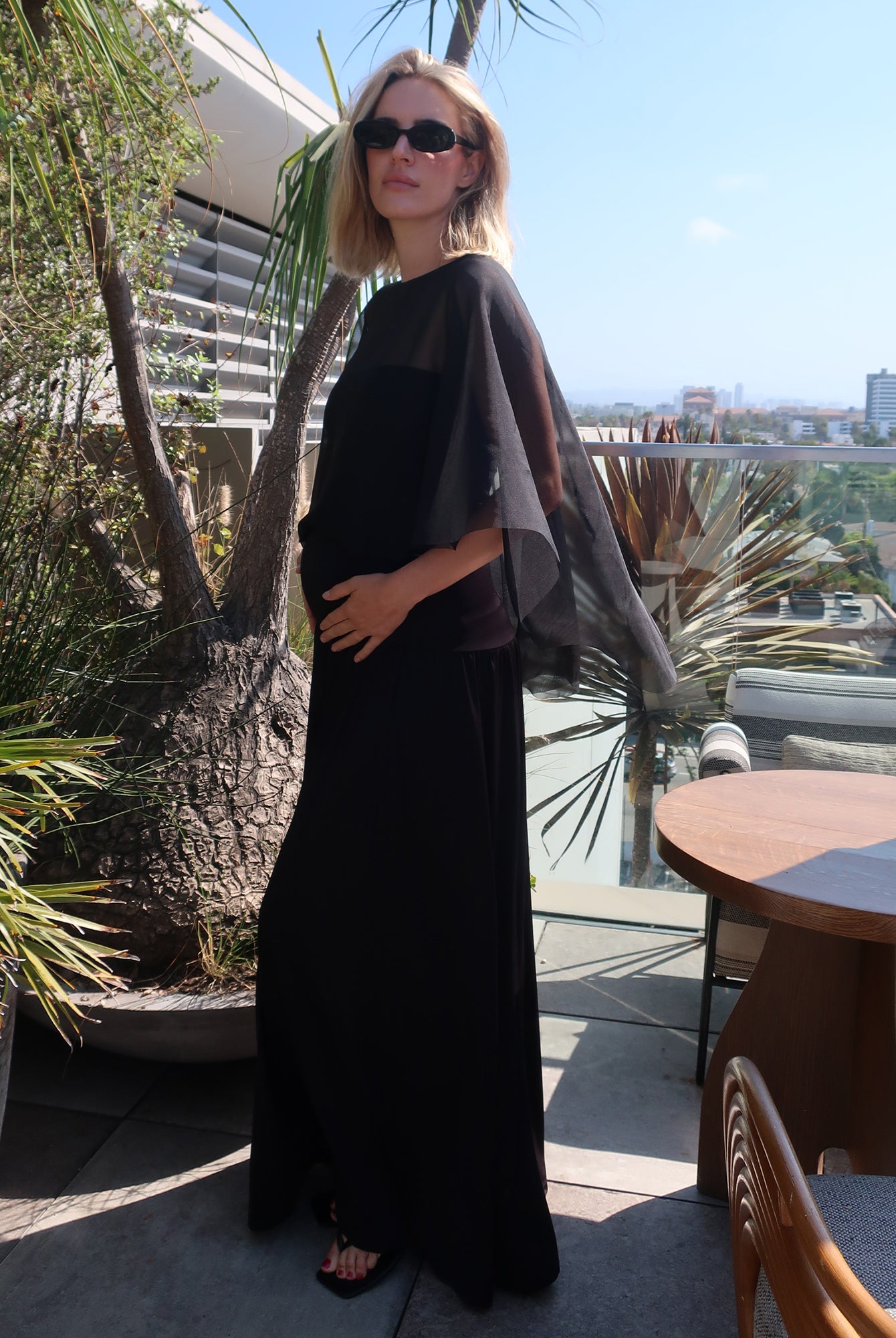 A woman wearing the black Bumpsuit Maria maxi dress with sheer chiffon cape and black sunglasses, standing on a sunny outdoor balcony with plants and a table in the background.