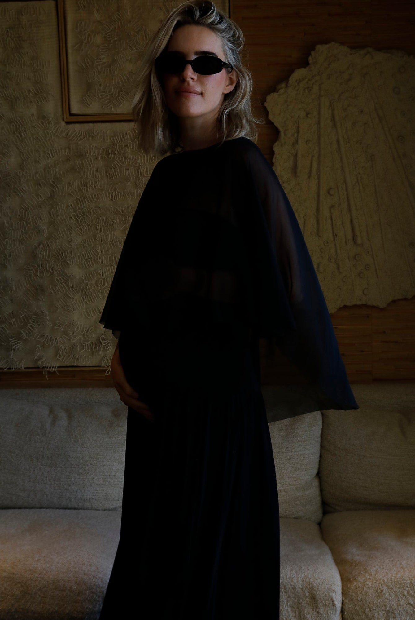 A woman wearing a Bumpsuit long black dress with a chiffon capulet and black sunglasses, standing in a dimly lit room with a textured beige sofa and wall art behind her.