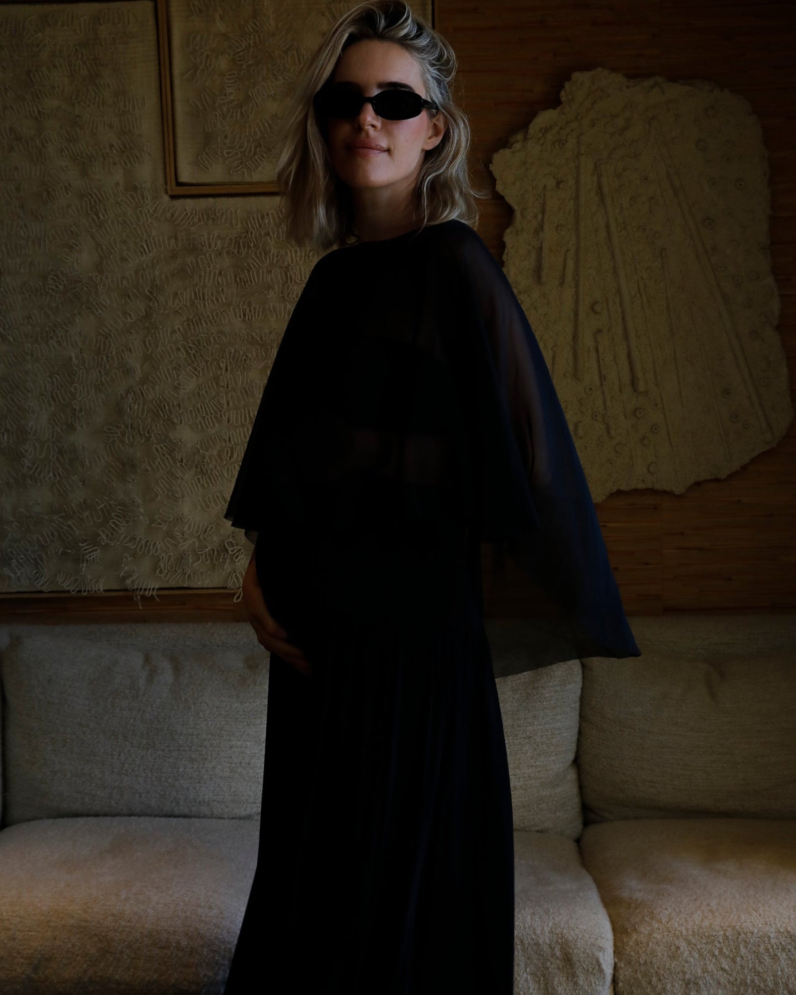 A woman wearing a Bumpsuit long black dress with a chiffon capulet and black sunglasses, standing in a dimly lit room with a textured beige sofa and wall art behind her.