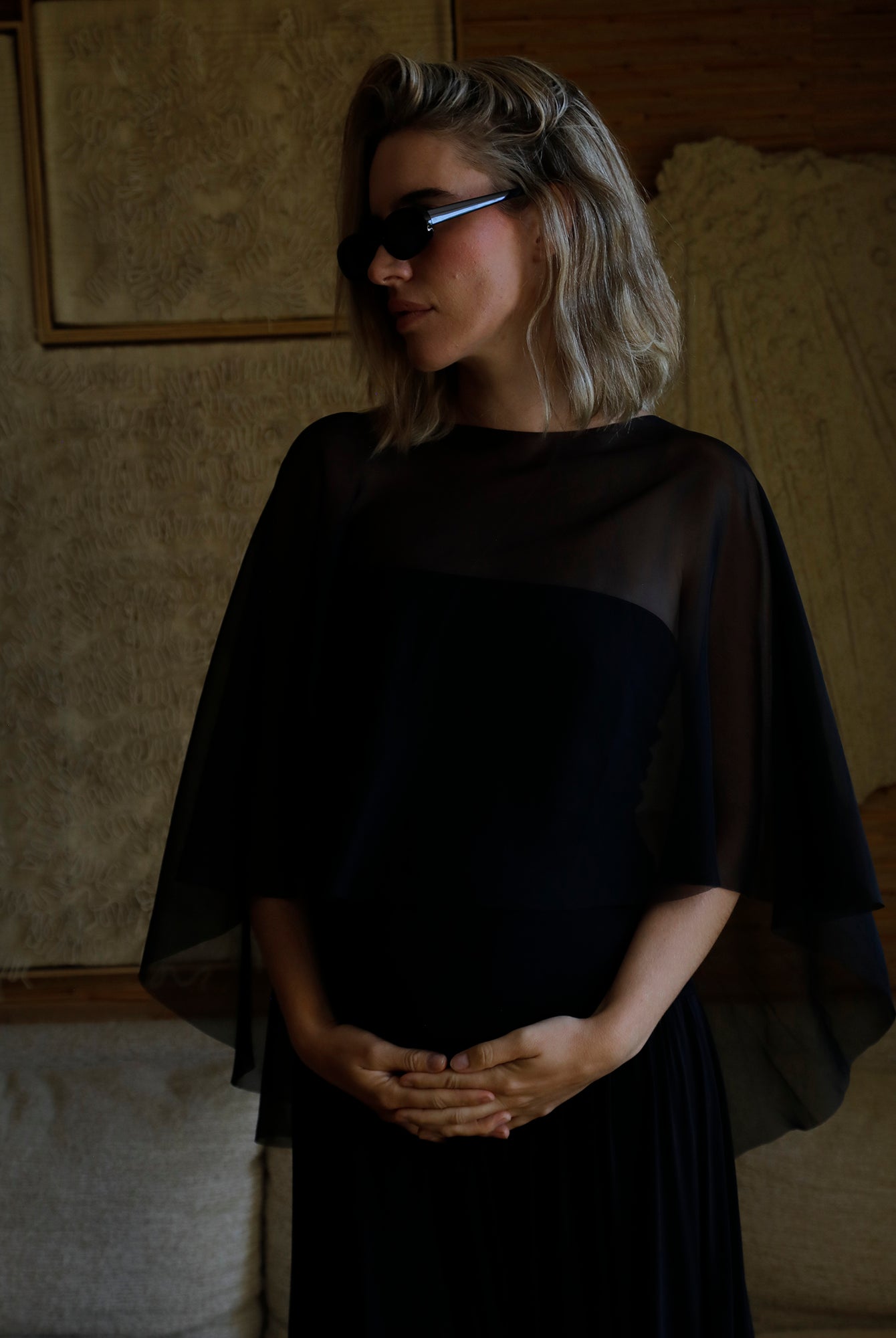 A woman with blonde hair wearing a black sheer cape-sleeve and dress by Bumpsuit, styled with black sunglasses, standing in a dimly lit room.