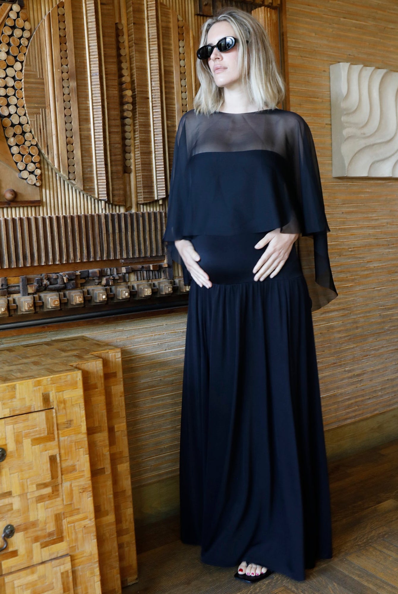 A pregnant woman wearing a long black maternity dress by Bumpsuit with a chiffon capulet and black sunglasses, standing in a modern room with wood-paneled walls and decorative wooden furniture.
