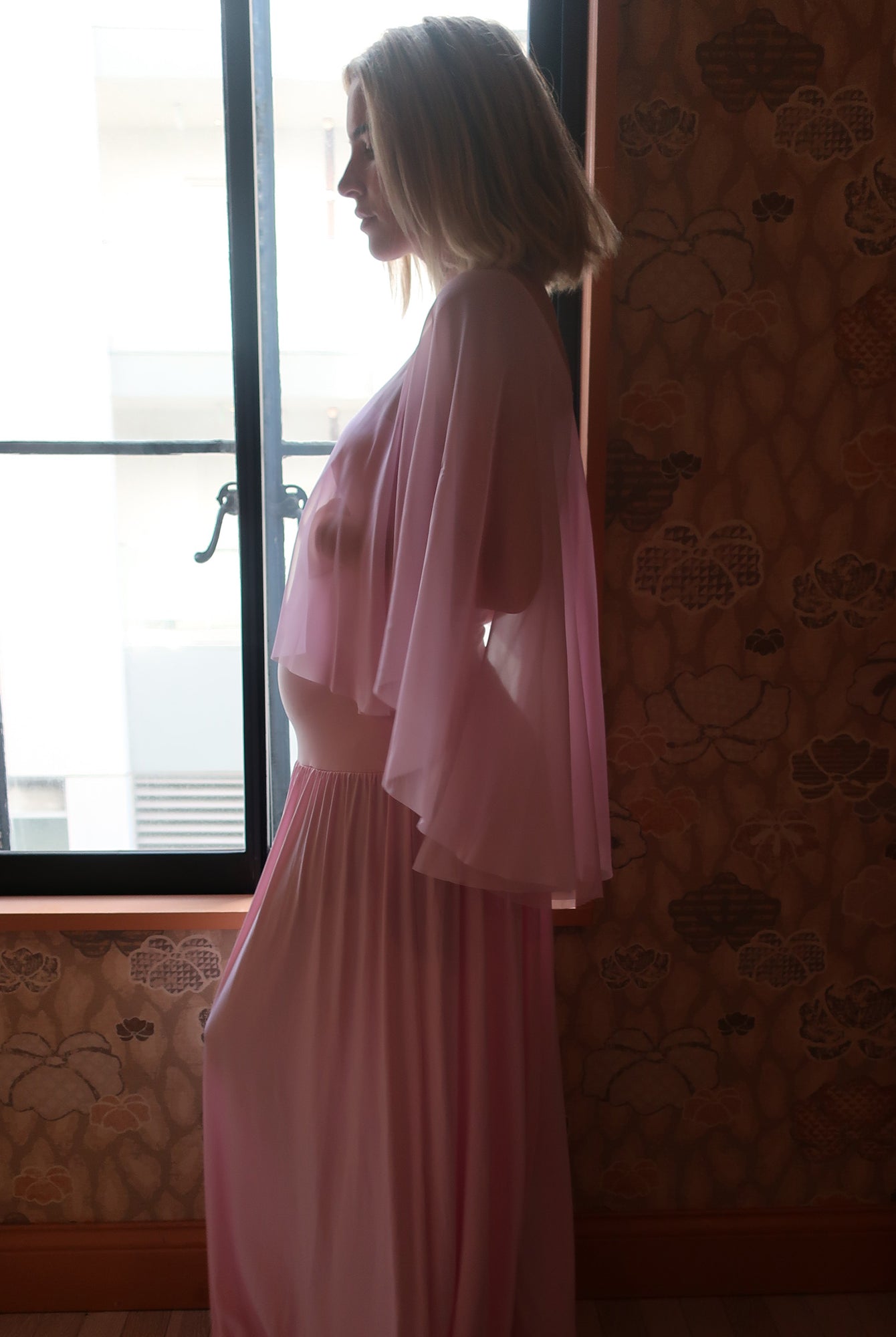 A pregnant woman in profile wearing a long pink dress by Bumpsuit with a sheer chiffon cape, standing indoors near a window with patterned wallpaper.
