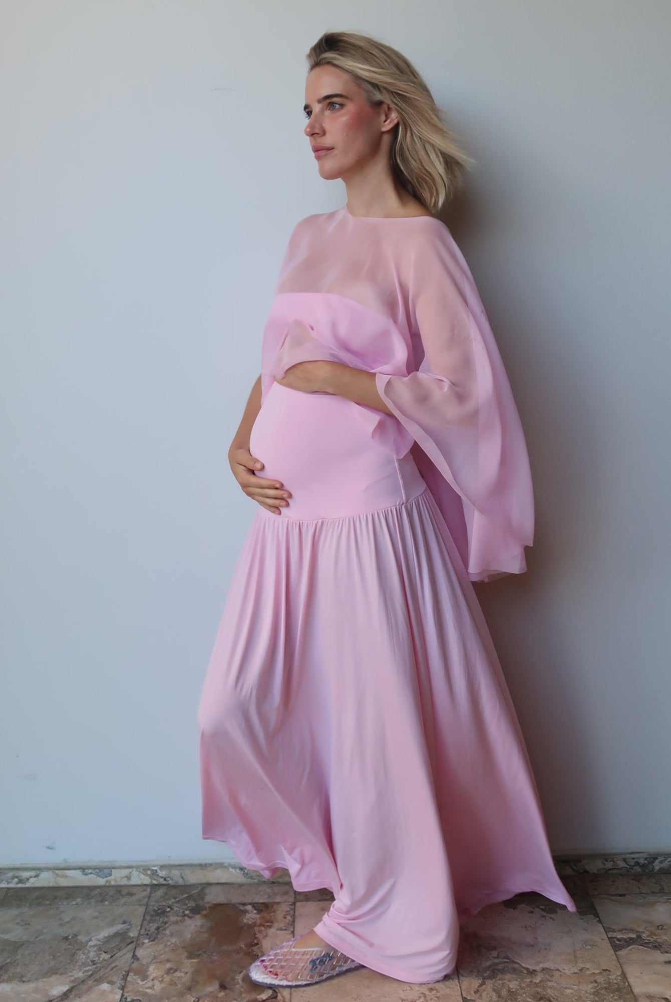 A pregnant woman wearing a light pink maternity dress with a sheer chiffon cape, standing against a plain light gray wall on a stone-tiled floor.