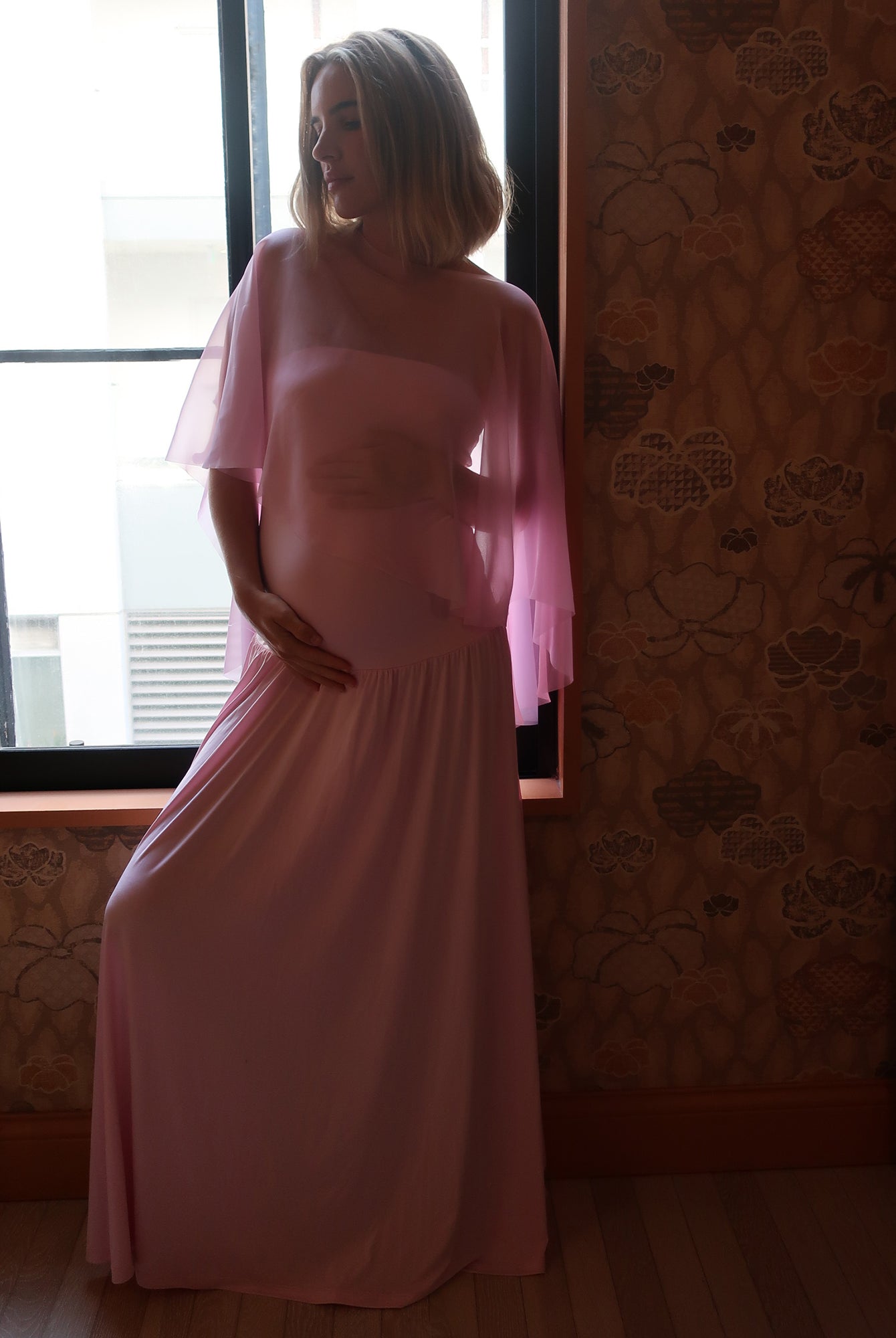A pregnant woman wearing a Bumpsuit long, flowing light pink gown with a chiffon cape stands in front of a window, with patterned wallpaper and wood flooring visible in the room.