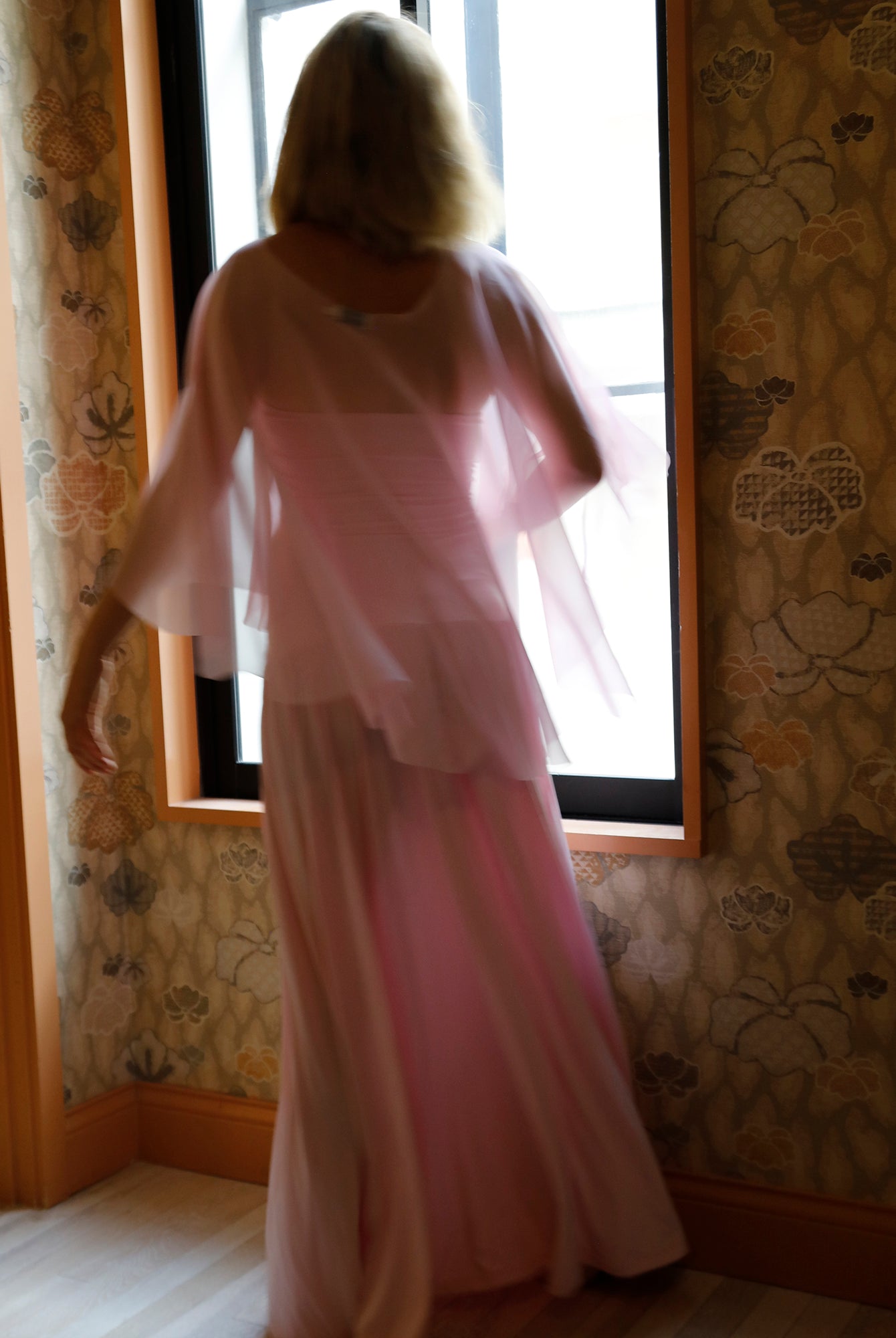 A woman viewed from behind wearing a light pink dress and chiffon cape by Bumpsuit, standing in front of a window in a room with patterned wallpaper.