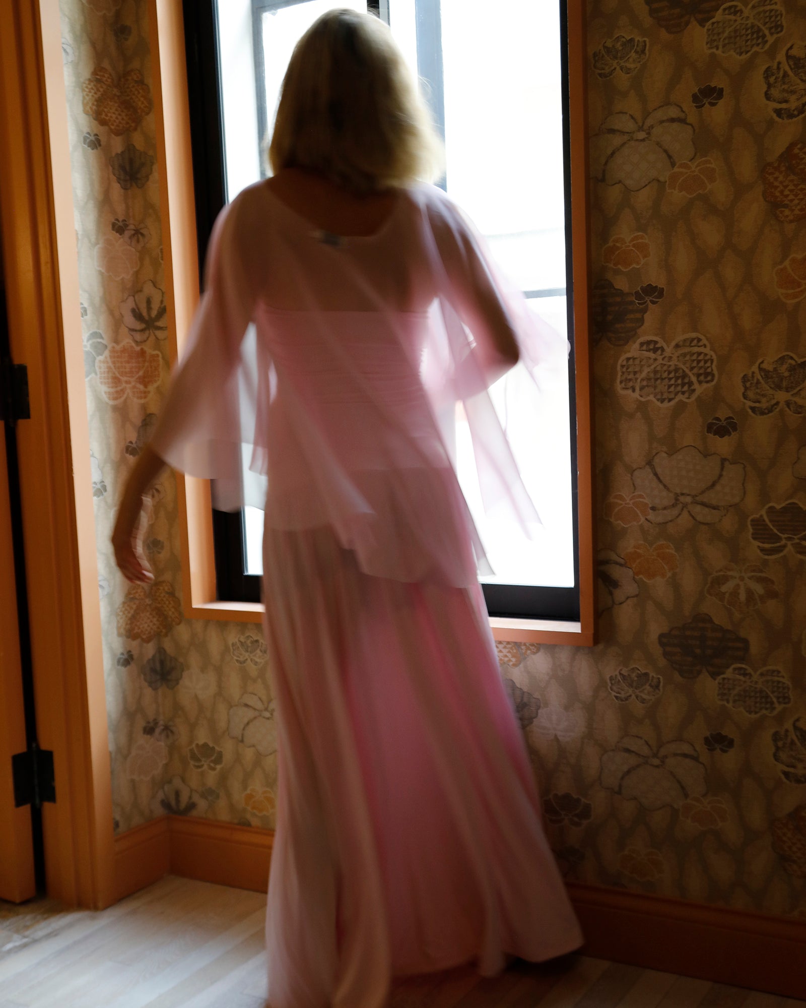 A woman viewed from behind wearing a light pink dress and chiffon cape by Bumpsuit, standing in front of a window in a room with patterned wallpaper.