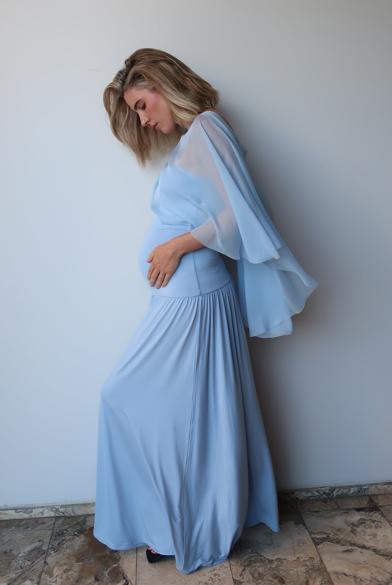 A pregnant woman wearing a light blue maternity maxi dress with a sheer chiffon cape, posing against a plain light gray wall.