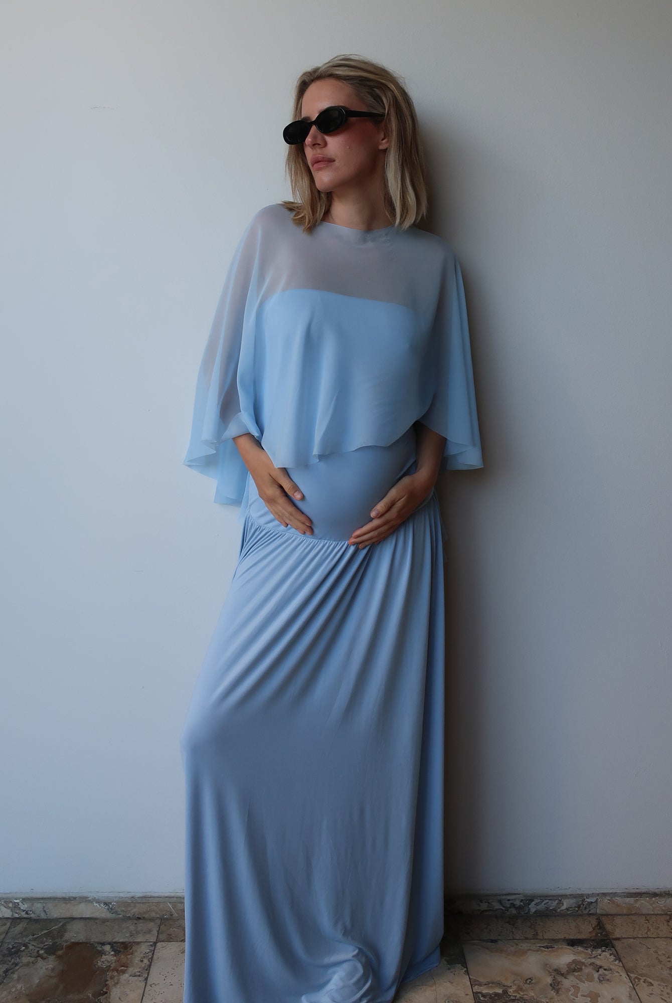 A pregnant woman wearing a Bumpsuit light blue maternity dress with a chiffon cape, standing against a plain light gray wall.