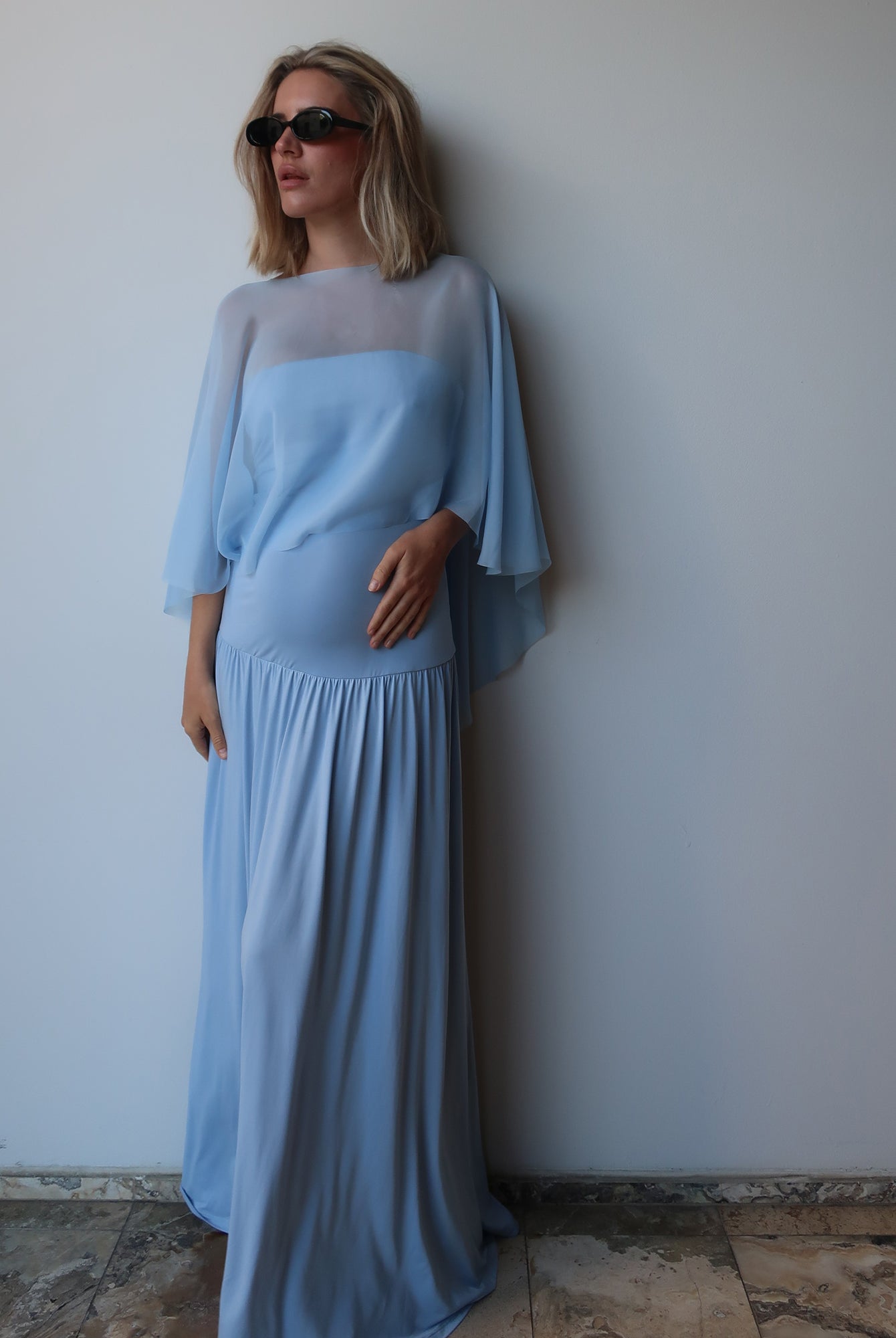 A pregnant woman wearing a light blue maxi dress with a chiffon cape, standing against a plain light gray wall.