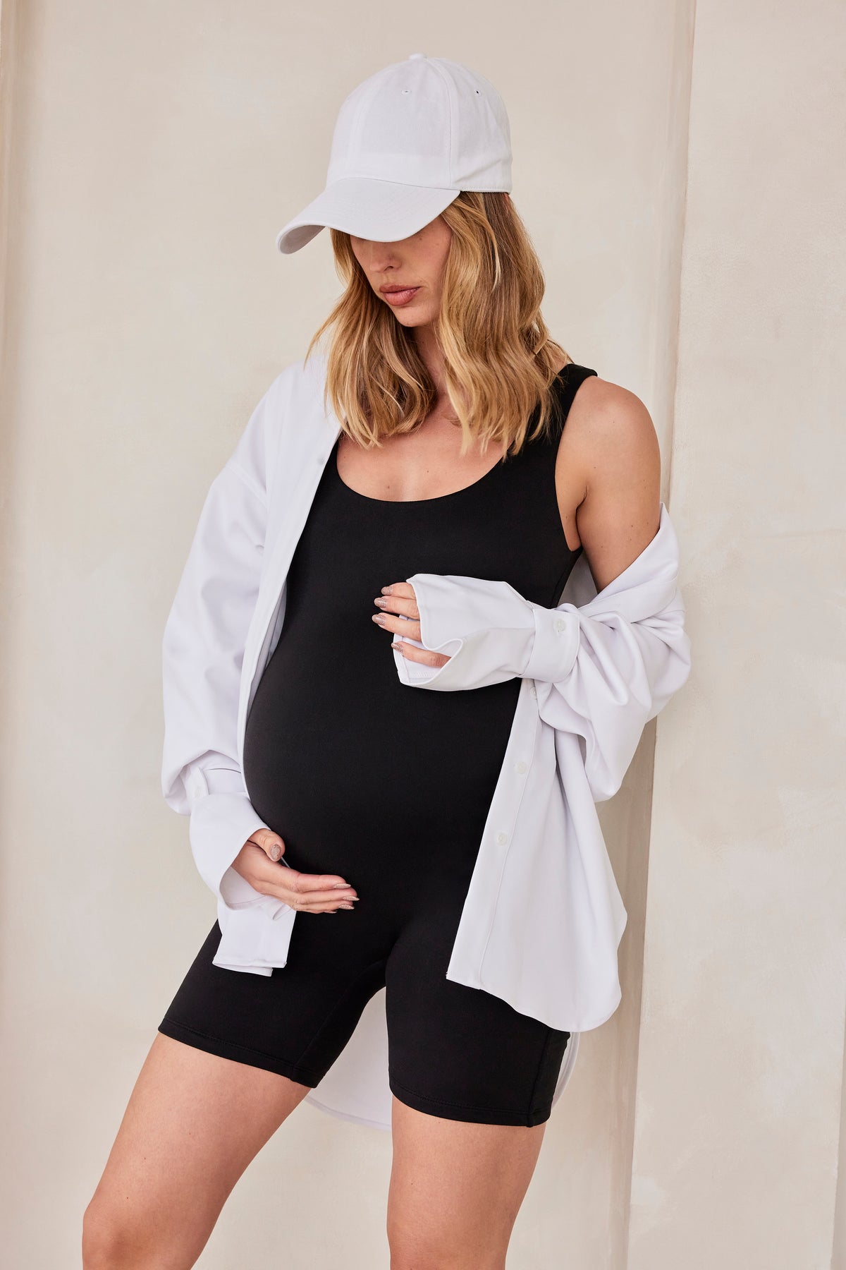 Pregnant woman wearing Bumpsuits scoop neck Cindy Romper and white shirt and hat against a feature wall