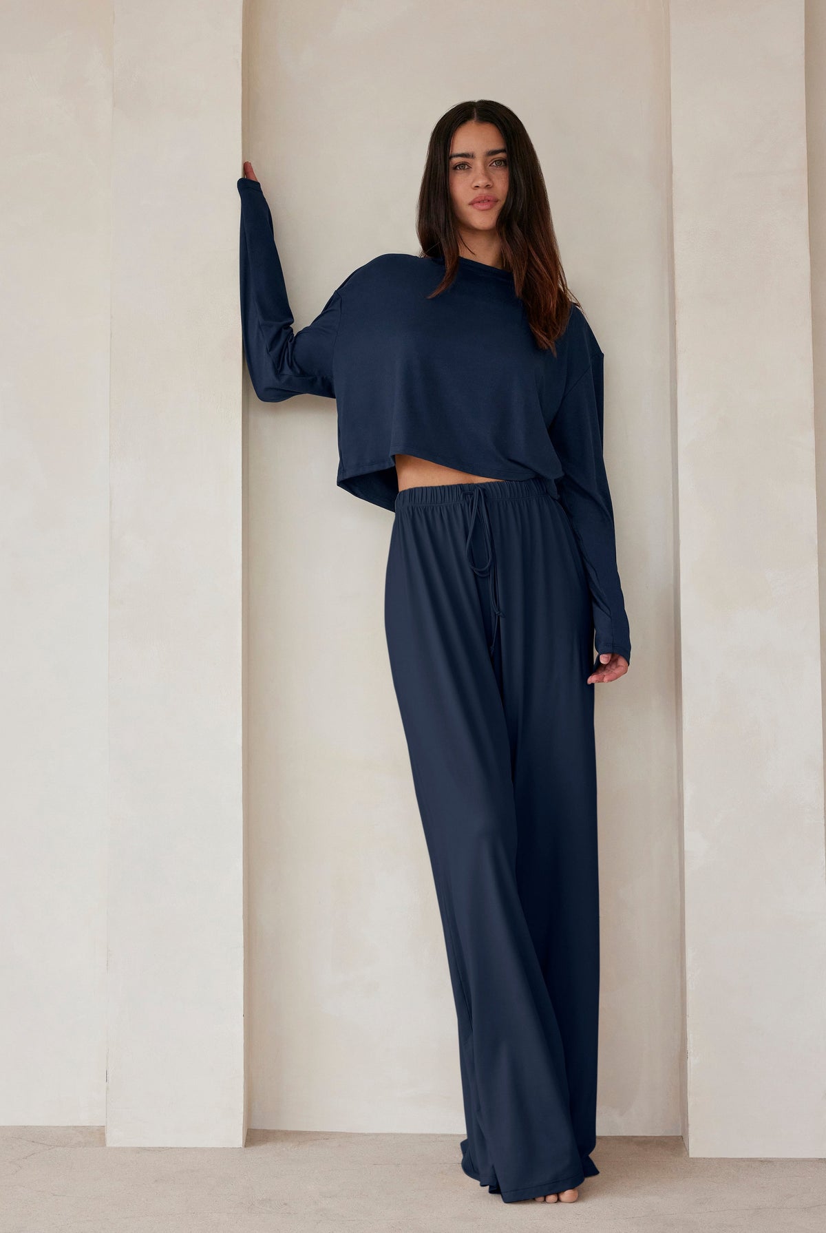 A woman standing against a beige wall in navy lounge pants and a crop long sleeve tee by Bumpsuit