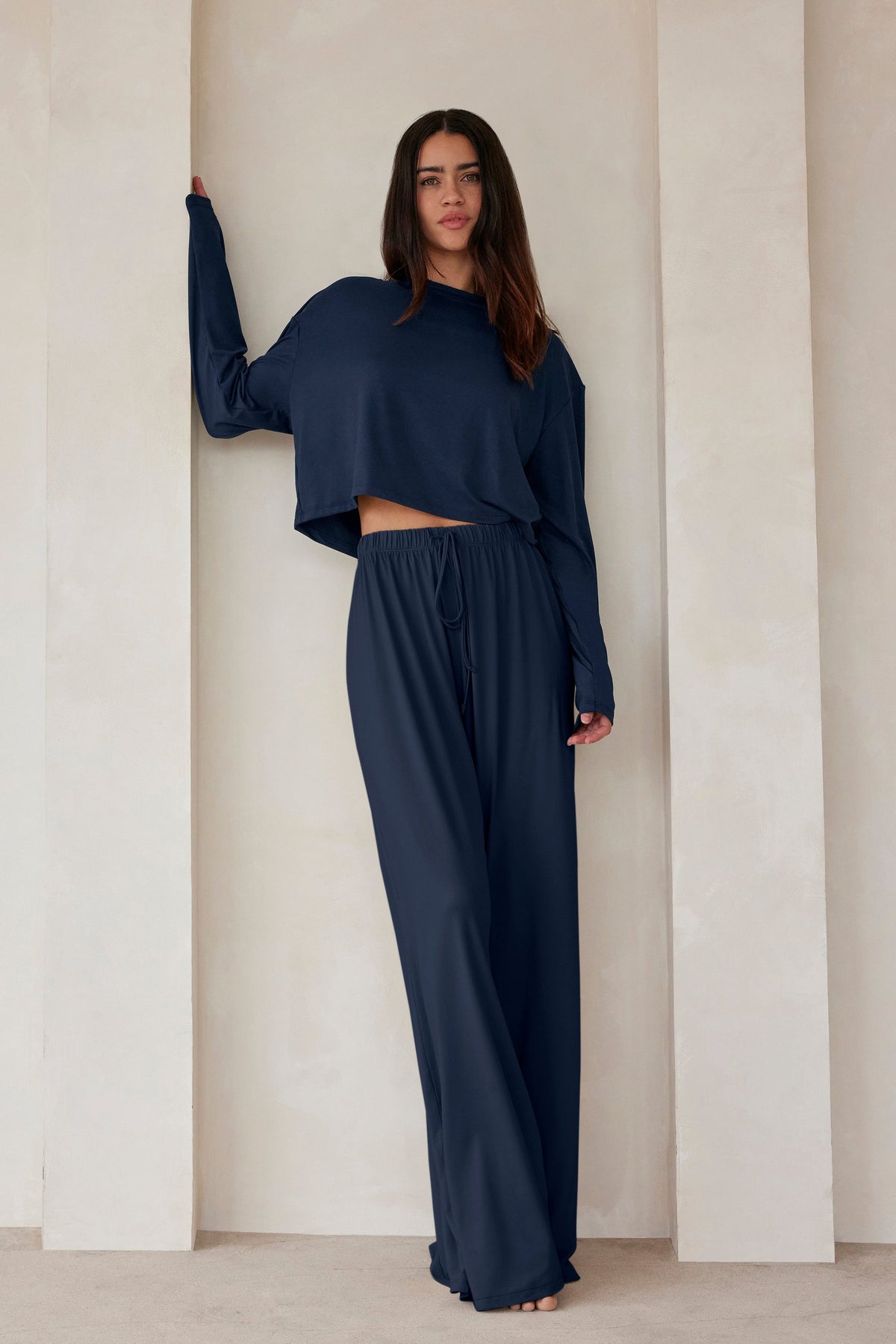 A woman standing against a beige wall in navy lounge pants and a crop long sleeve tee by Bumpsuit