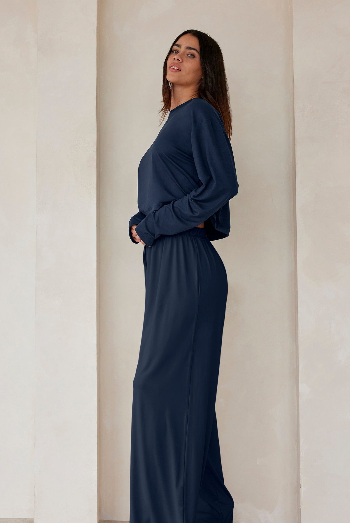 A woman standing against a beige feature wall wearing Bumpsuits Cloud lounge pants and a long sleeve crop tee in navy