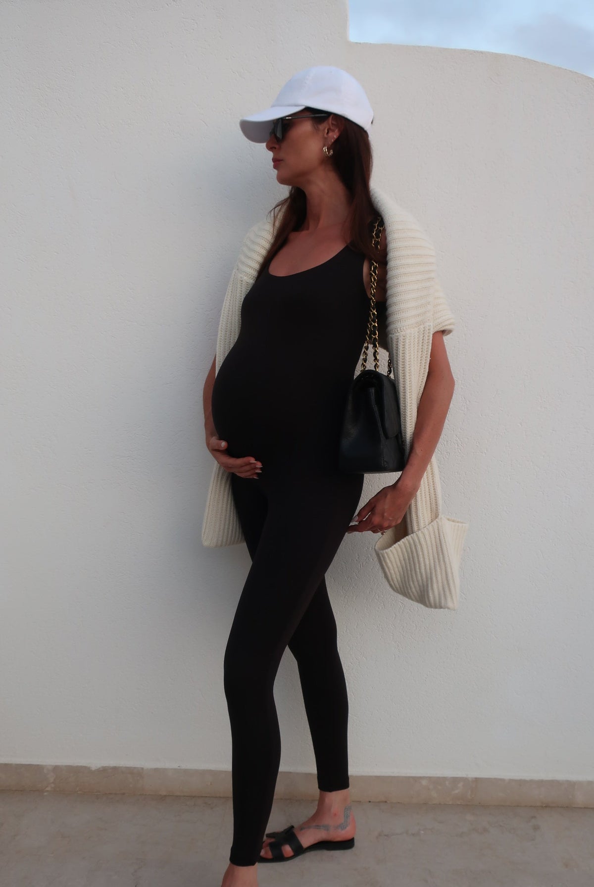Pregnant woman in black outfit with white cap sitting on a white chair against a light grey wall.