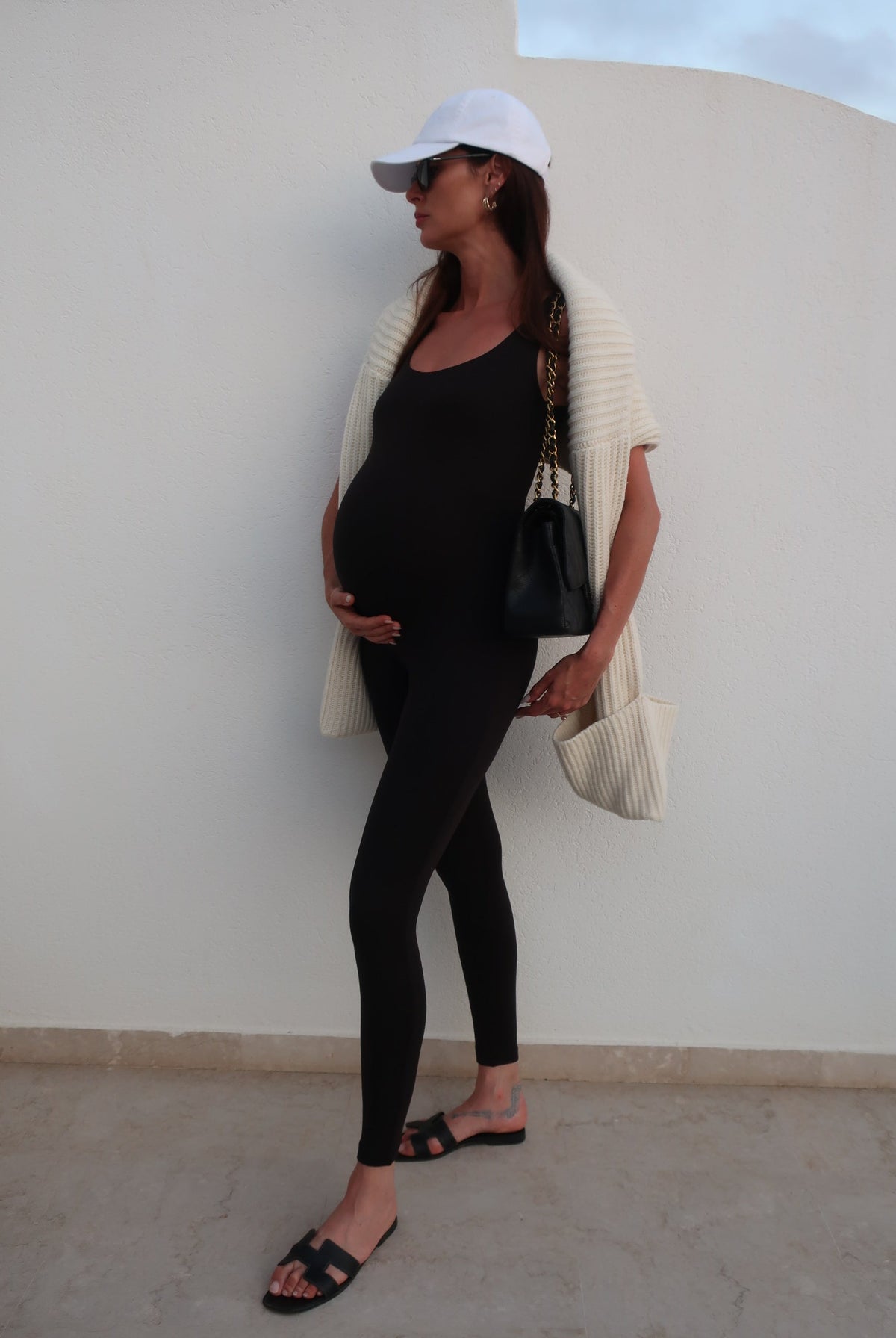 Pregnant woman in black outfit with white cap sitting on a white chair against a light grey wall.