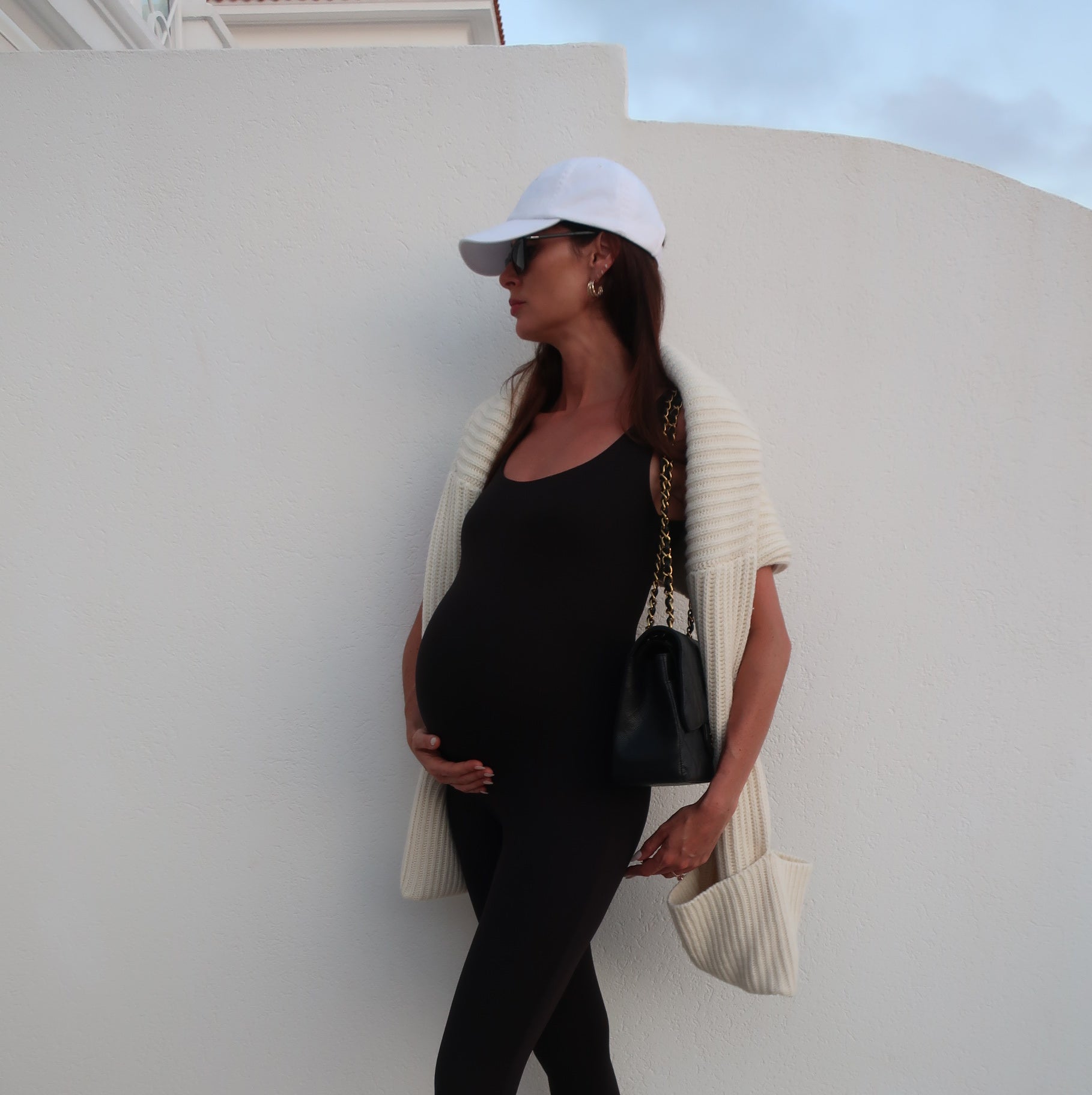 Pregnant woman in black outfit with white cap sitting on a white chair against a light grey wall.
