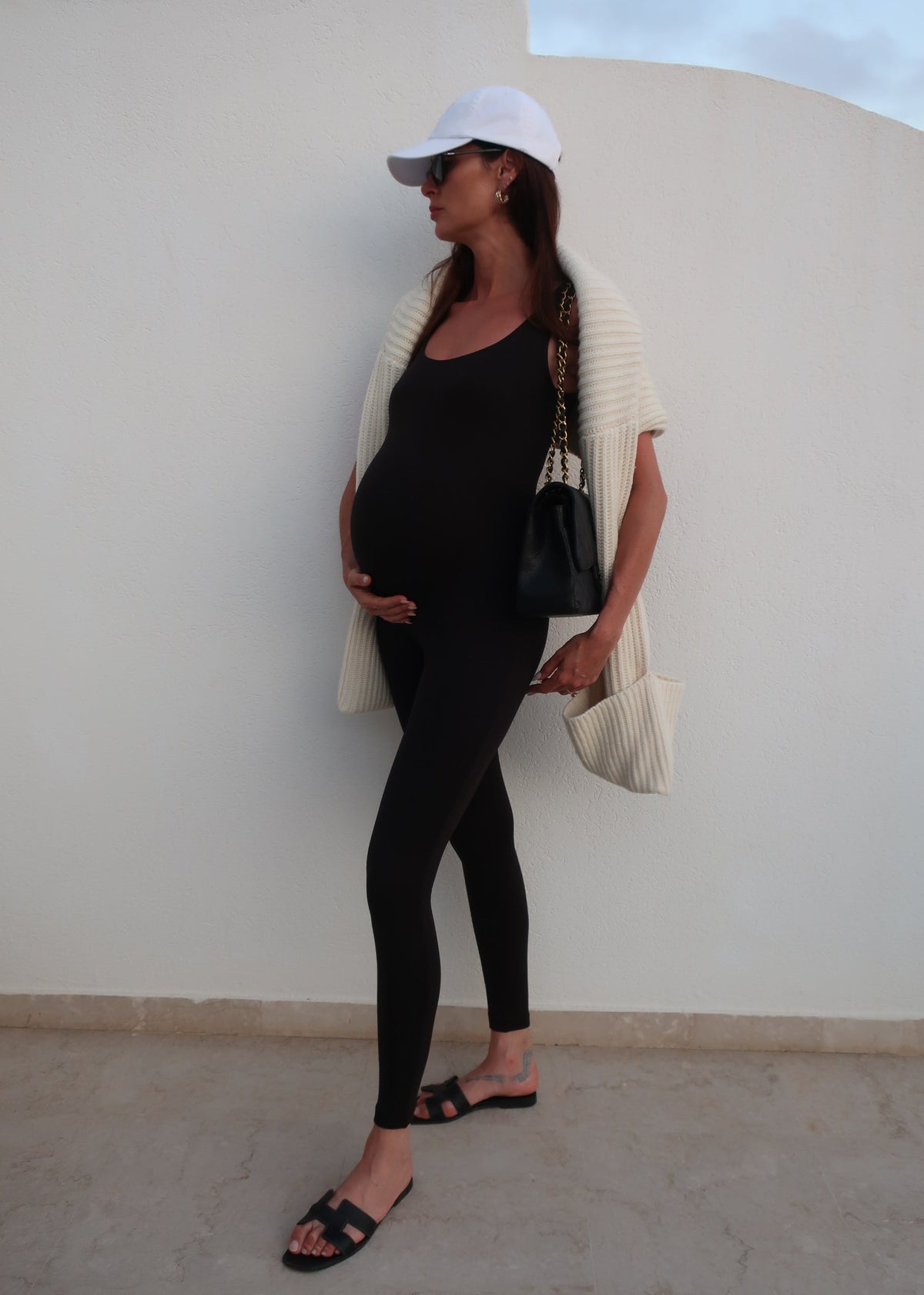 Pregnant woman in black outfit with white cap sitting on a white chair against a light grey wall.